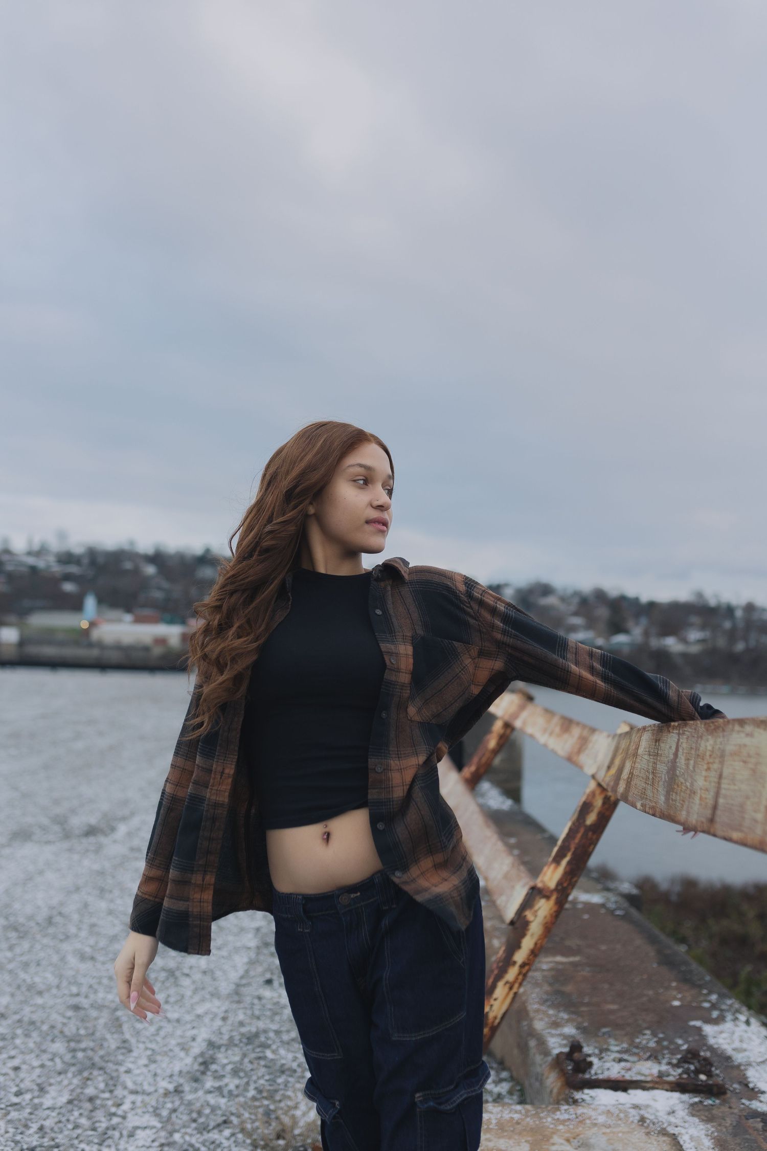 Person in plaid jacket and black crop top standing near waterfront on cloudy day.