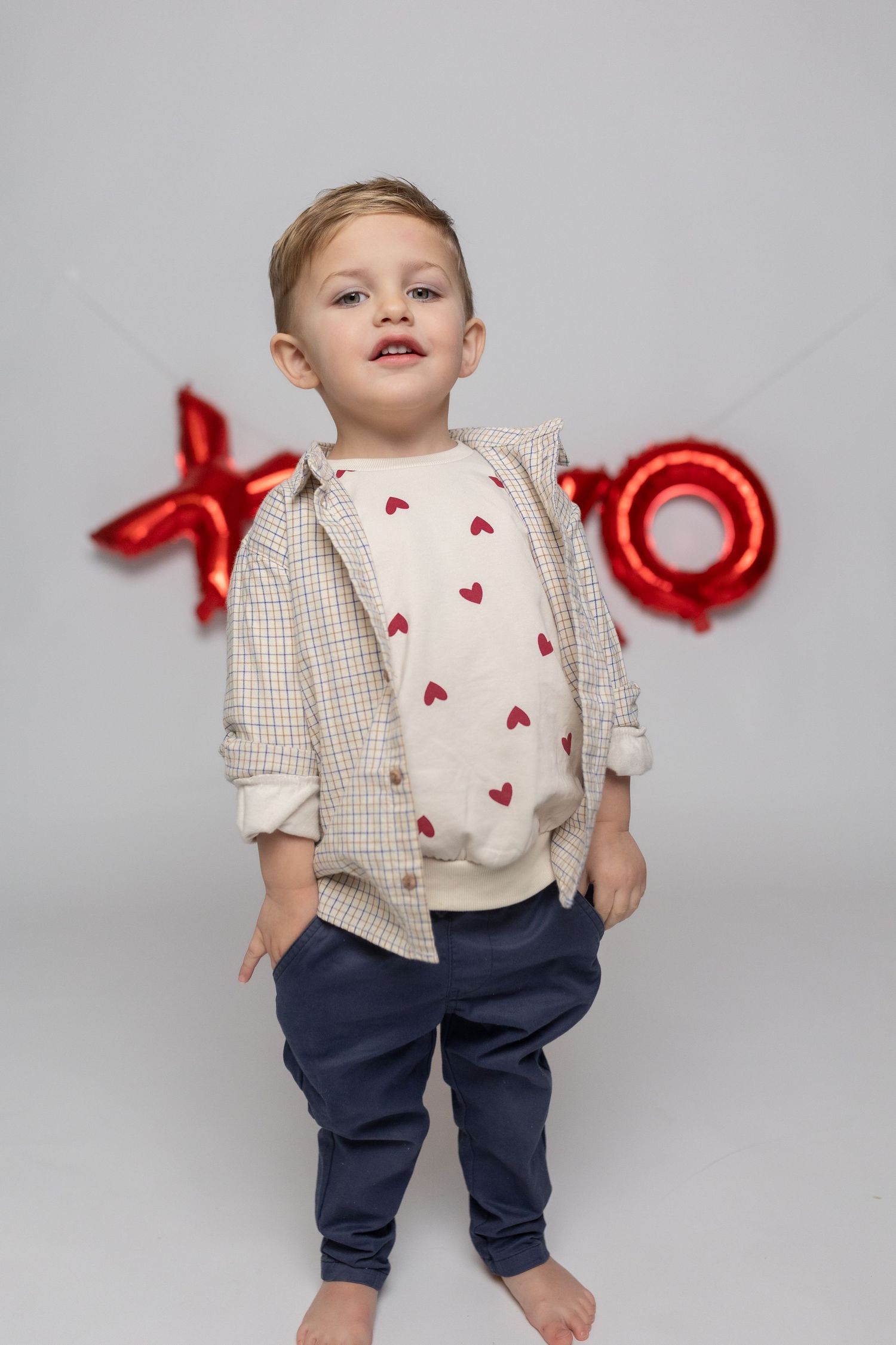 A child wearing heart-patterned shirt and plaid button-up against red balloon letters.