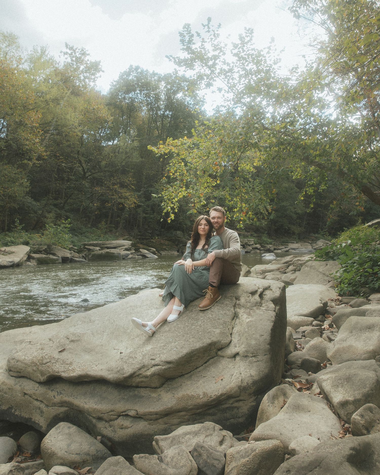 Three people relaxing on large gray rocks along a scenic creek surrounded by autumn trees and foliage.