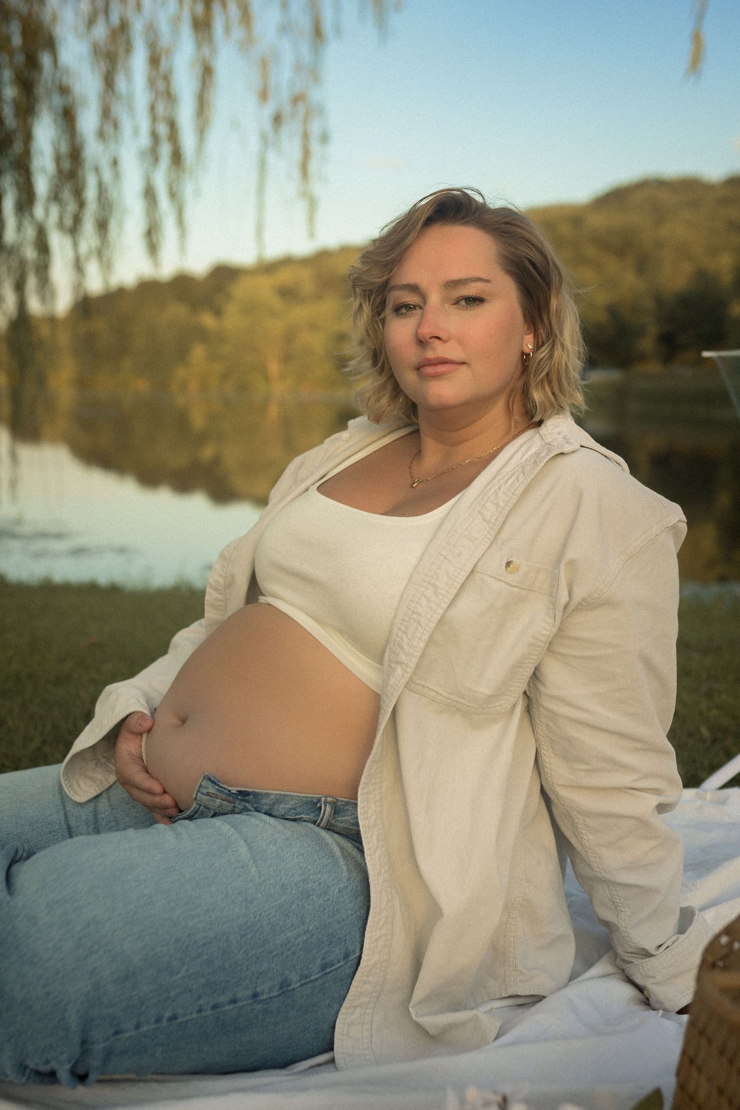 Person in white top and denim sits by lake at sunset.
