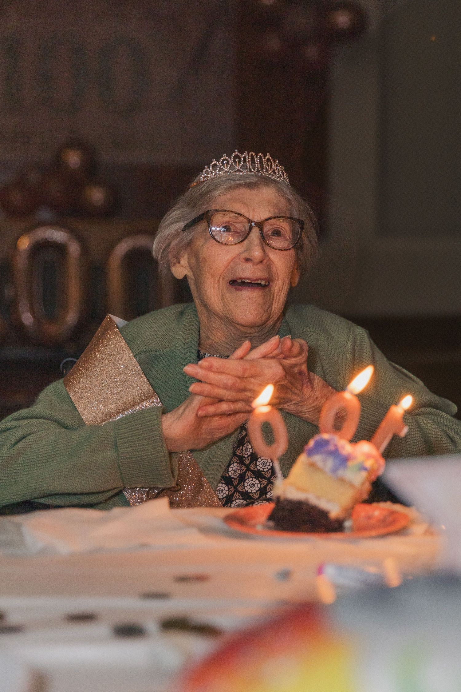 Elderly person wearing a tiara celebrating with lit birthday cake candles.