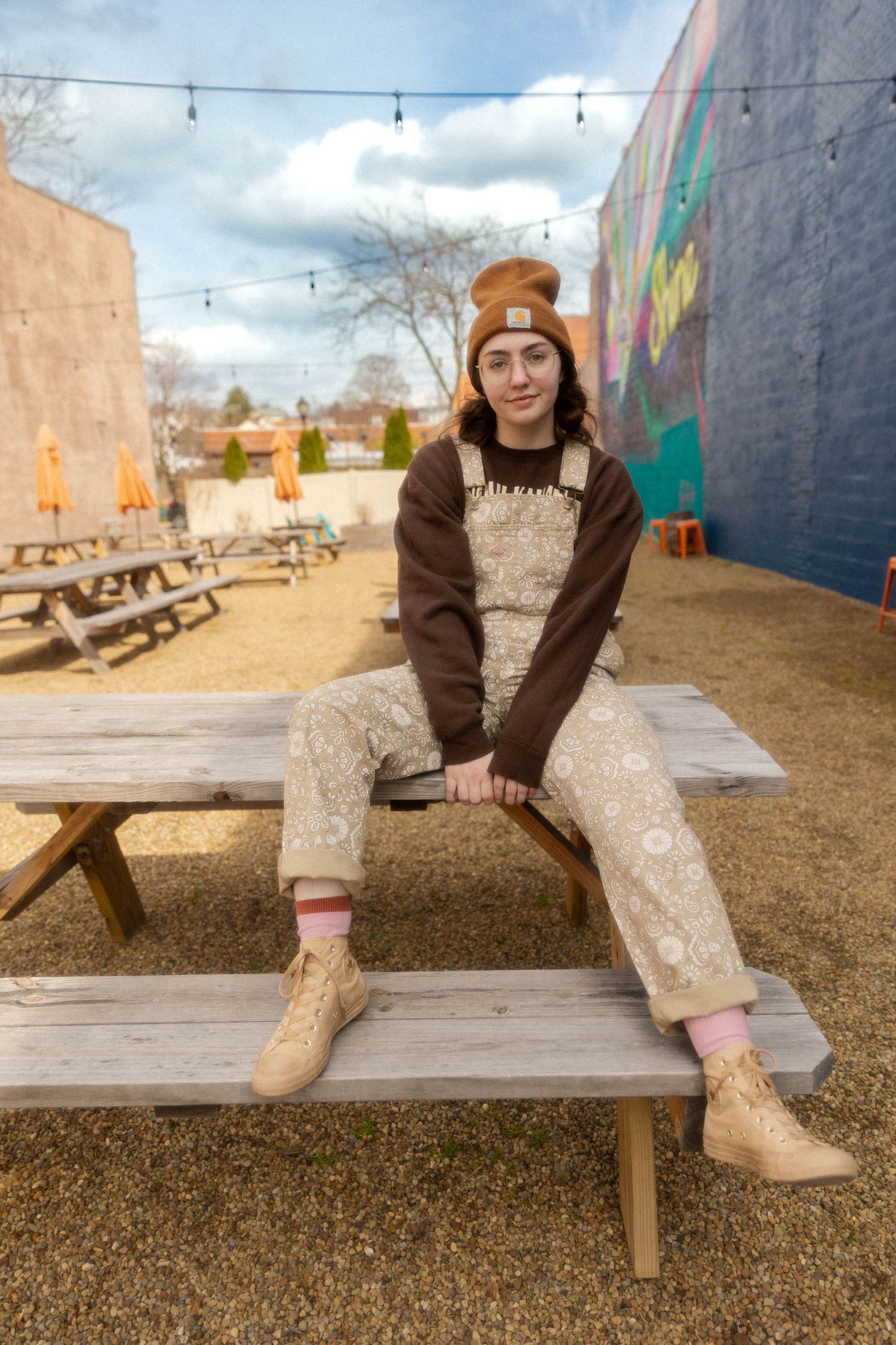 Casual outfit with brown beanie and overalls sitting on wooden picnic table in outdoor patio.