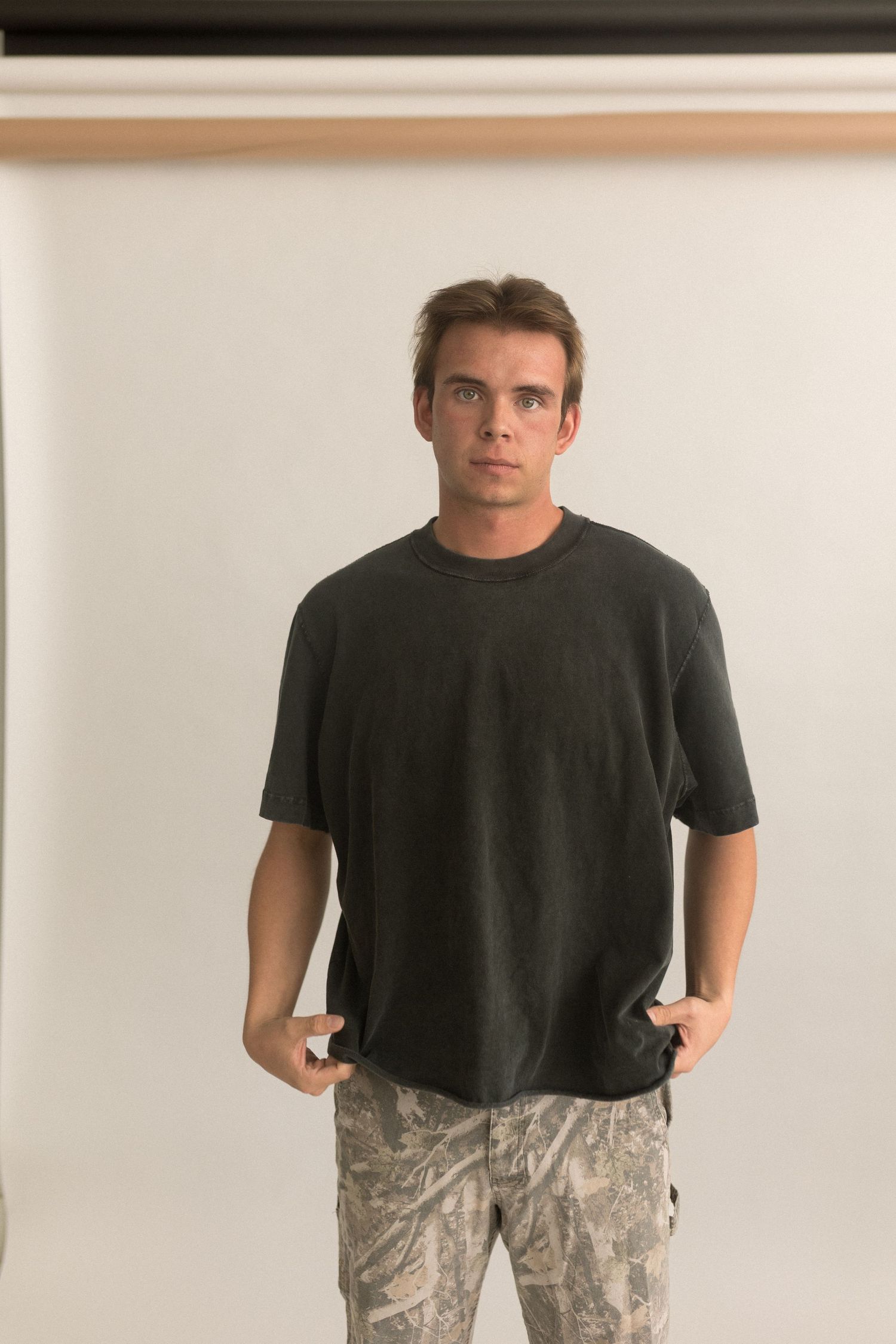 Person poses with hands in pockets wearing black t-shirt and camo pants against white backdrop.