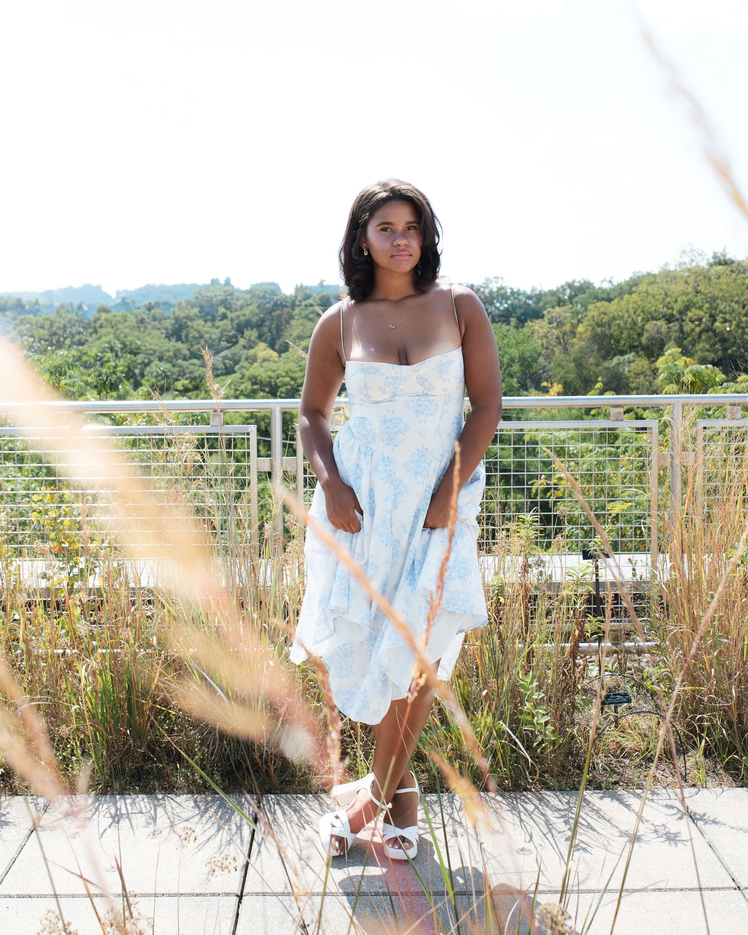 A person in a white summer dress poses on a wooden deck with tall grasses and greenery in the background.