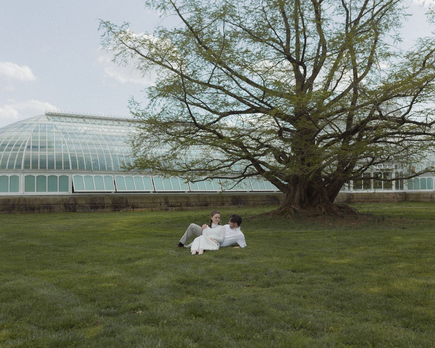 Couple relaxing on green grass near a large tree beside a glass greenhouse conservatory building.