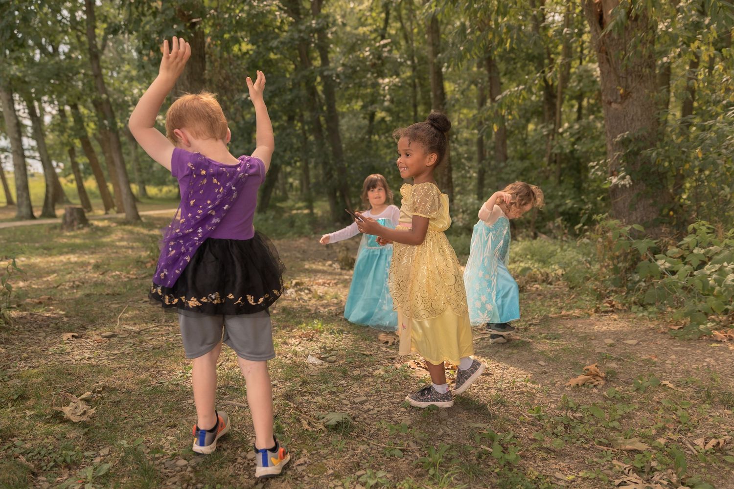 Children in princess dresses play and dance together in a wooded setting.