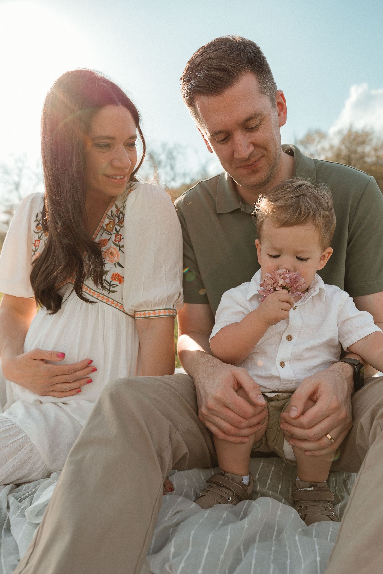A young family sits together outdoors on a sunny day while their toddler makes a silly face.