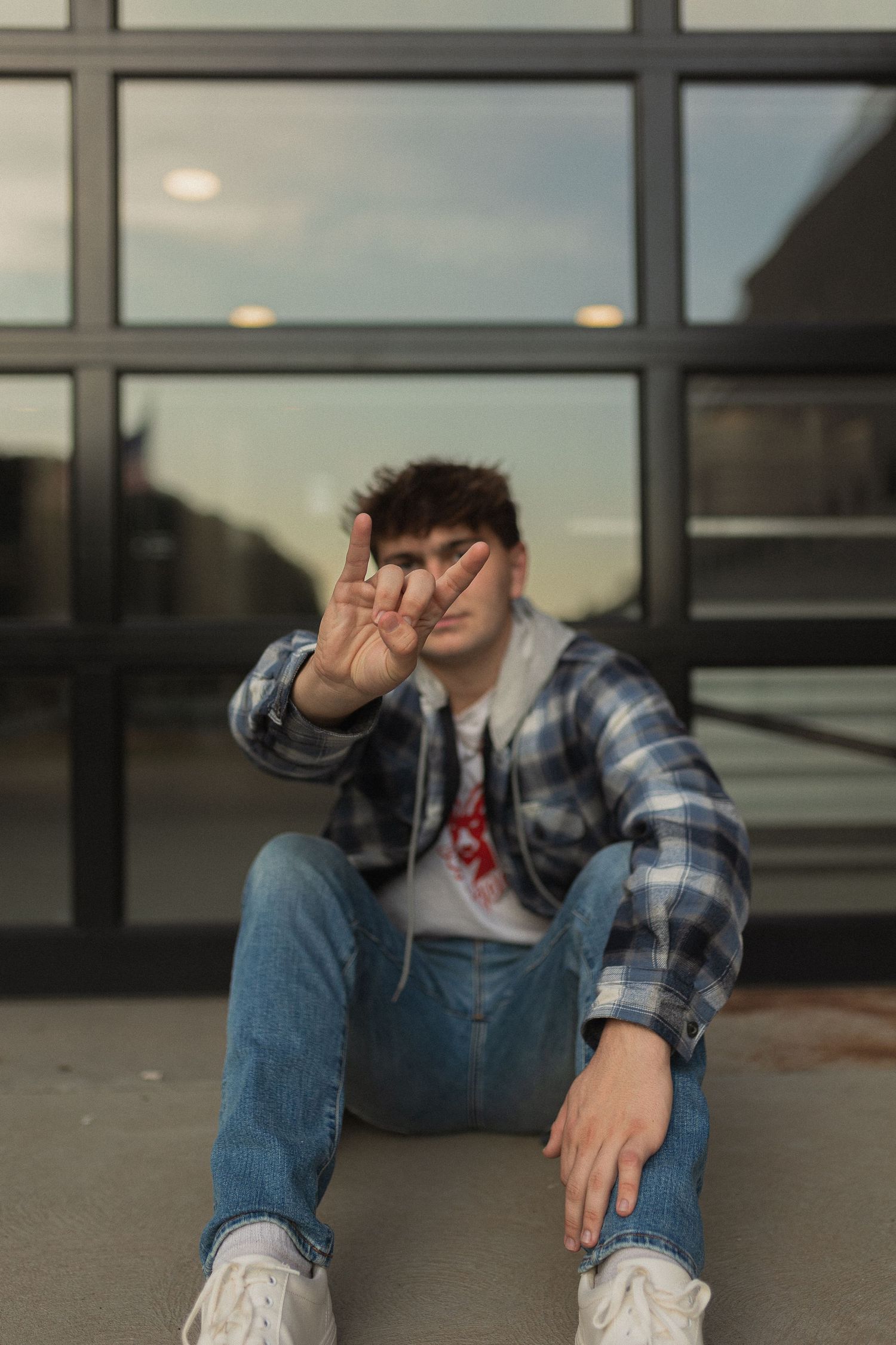 Person in casual wear making peace sign while sitting on ground near glass building.