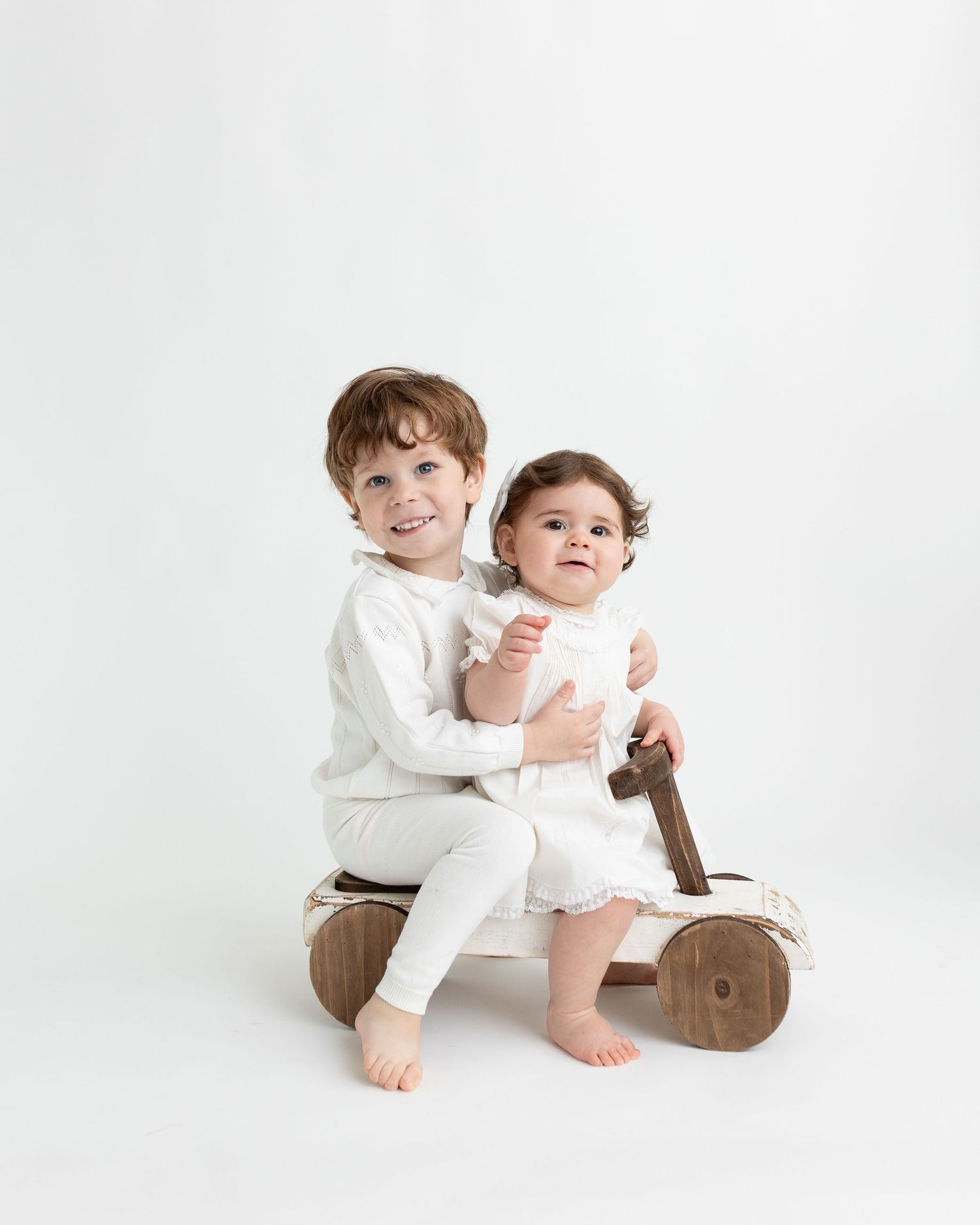 Two children in white outfits sit together on a wooden toy motorcycle against a white background.