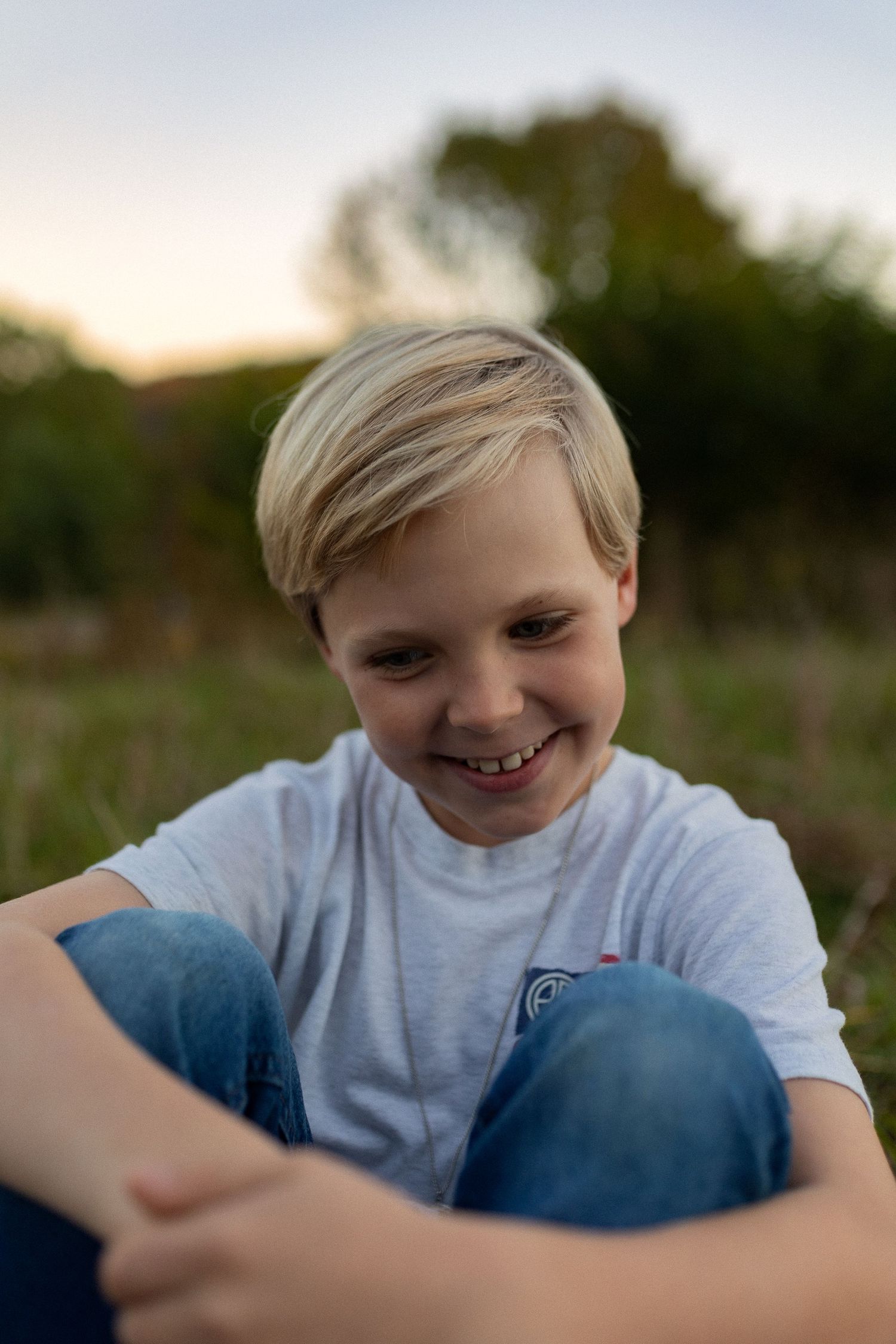Young person in gray t-shirt smiling while sitting outdoors at sunset.