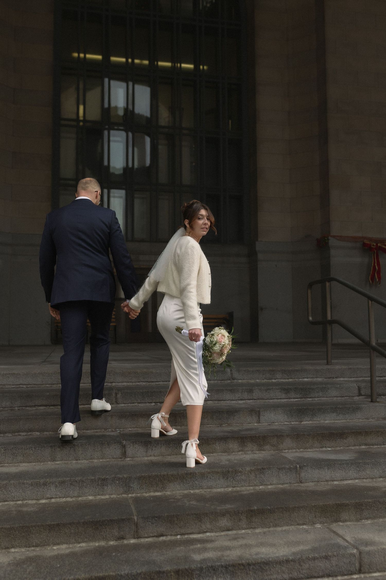 Couple ascending stone steps outside building, bride in modern short wedding dress.
