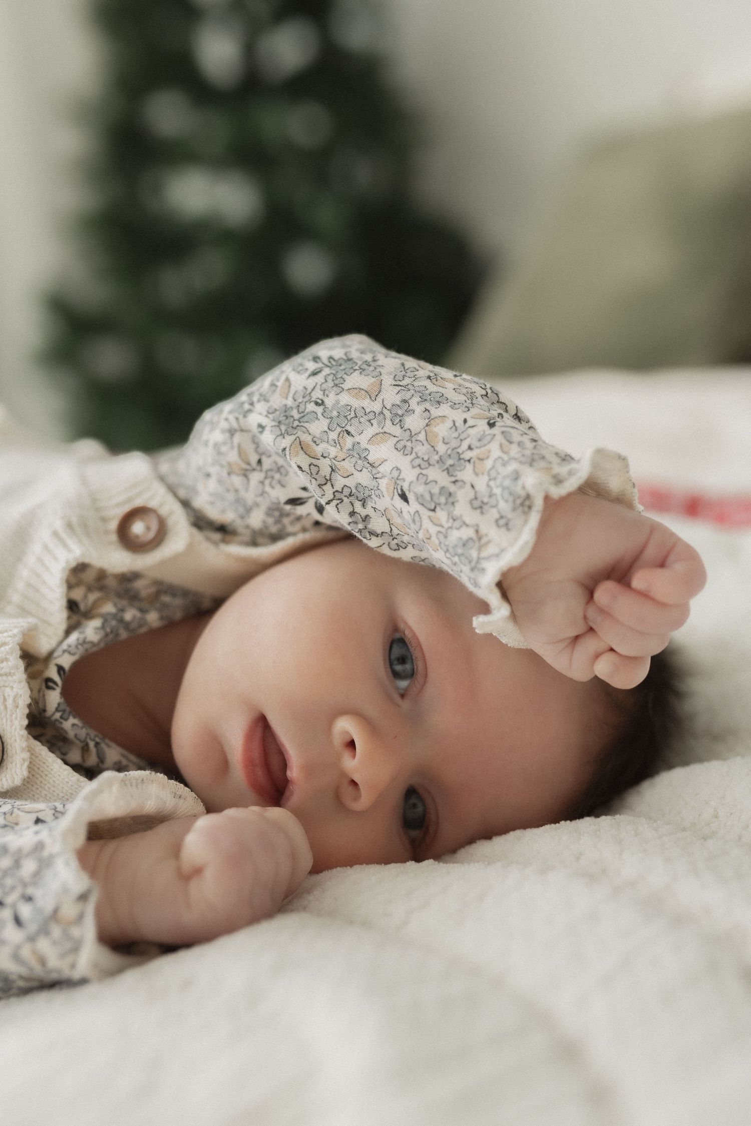 Baby in lace outfit lying on white bedding with Christmas tree in background.