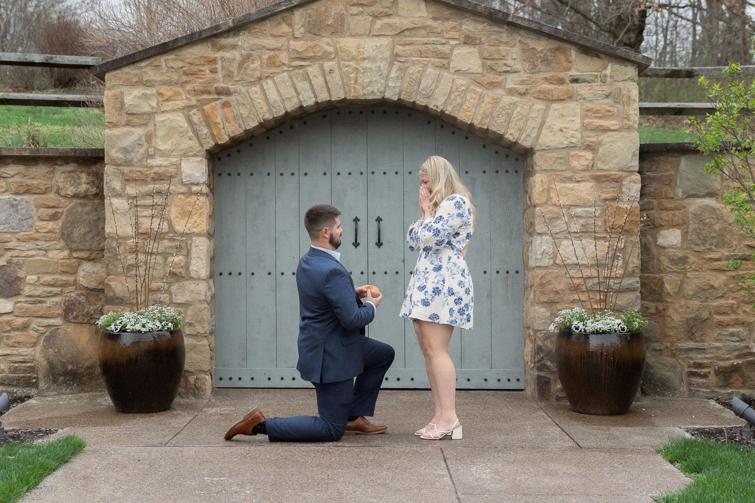 Surprise proposal captured outside a rustic stone arch, dreamy spring engagement with romantic storytelling, filmy-like and natural tones.