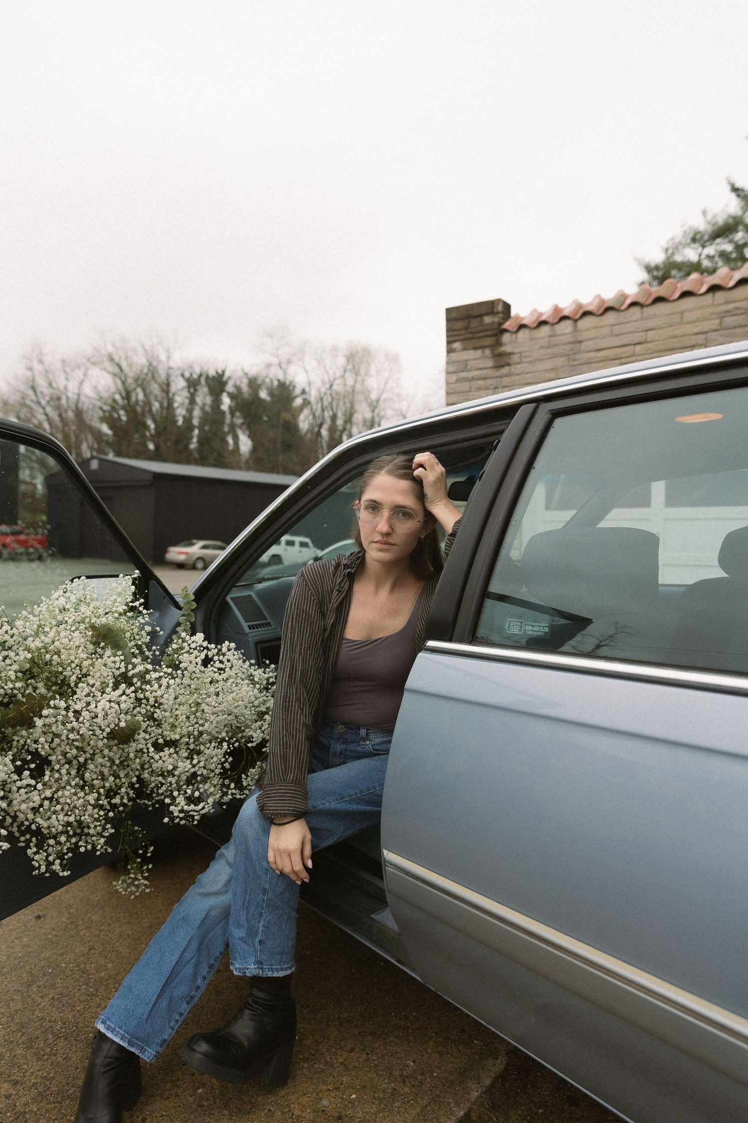 Someone sits in blue minivan with door open surrounded by baby's breath flowers in moody outdoor setting.