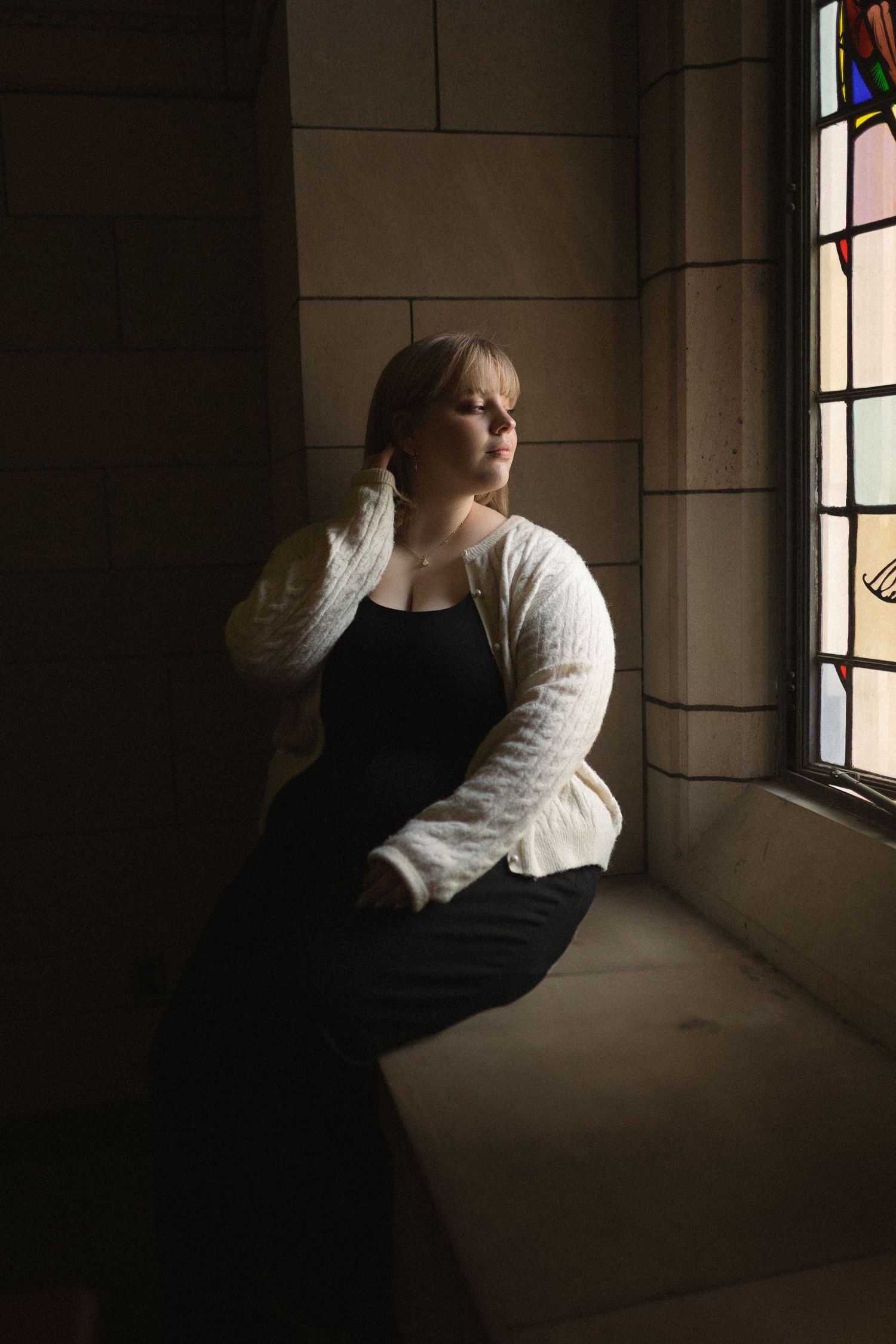 A contemplative figure in black dress and white cardigan sits near a stained glass window in moody church lighting.