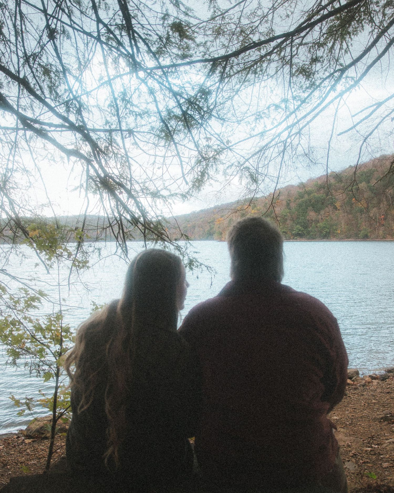 Silhouettes of people sitting by a serene lake surrounded by autumn trees and branches on a misty day.