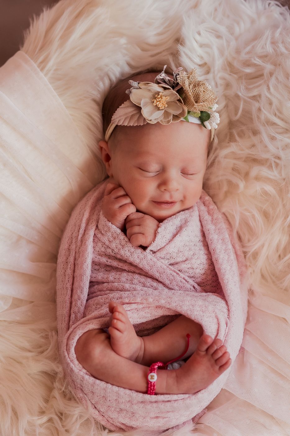 Newborn baby girl wrapped in soft pink blanket with floral headband, posed on fur in natural light newborn photography.