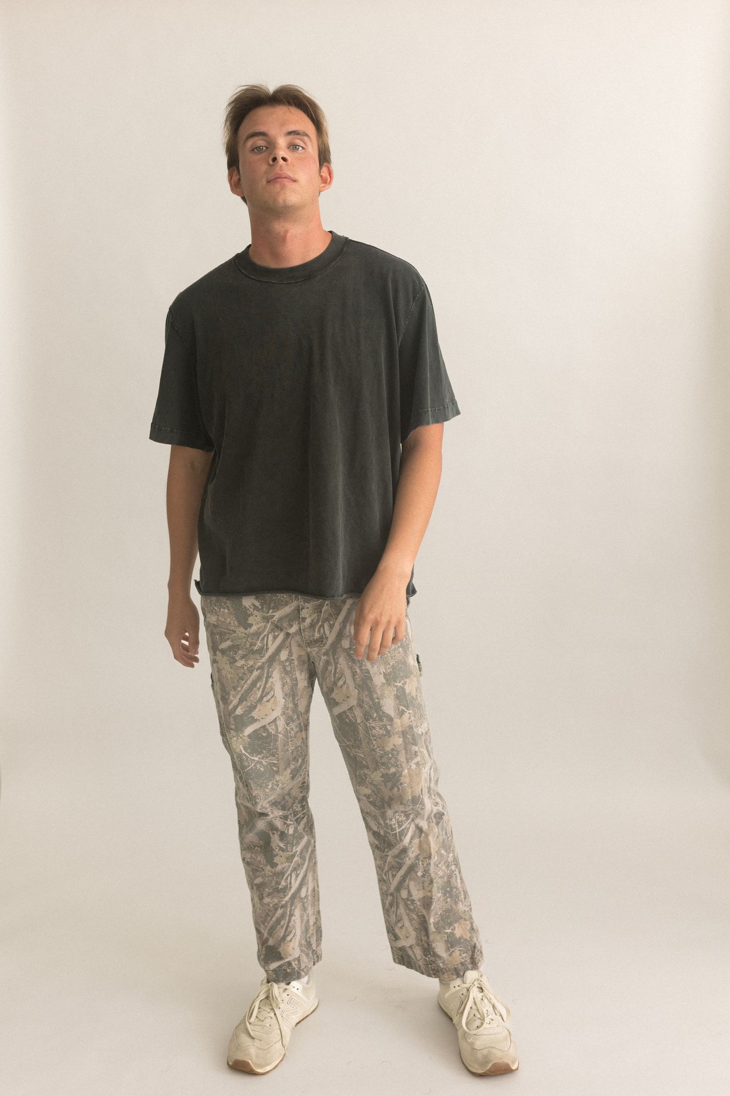 Person stands confidently in oversized black tee and camo pants against white background.