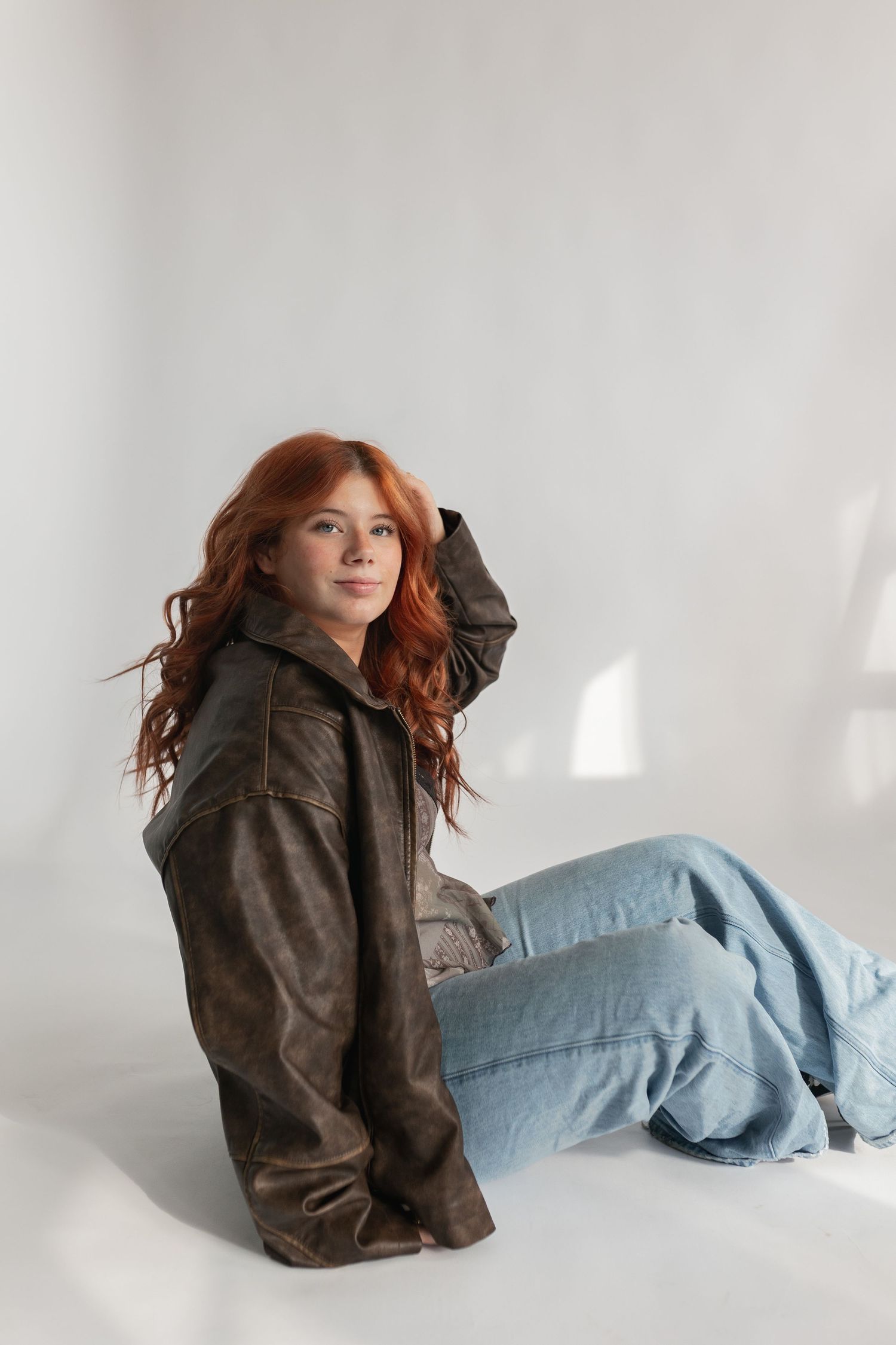 Fashion shot of redhead in oversized leather jacket and denim against white backdrop.