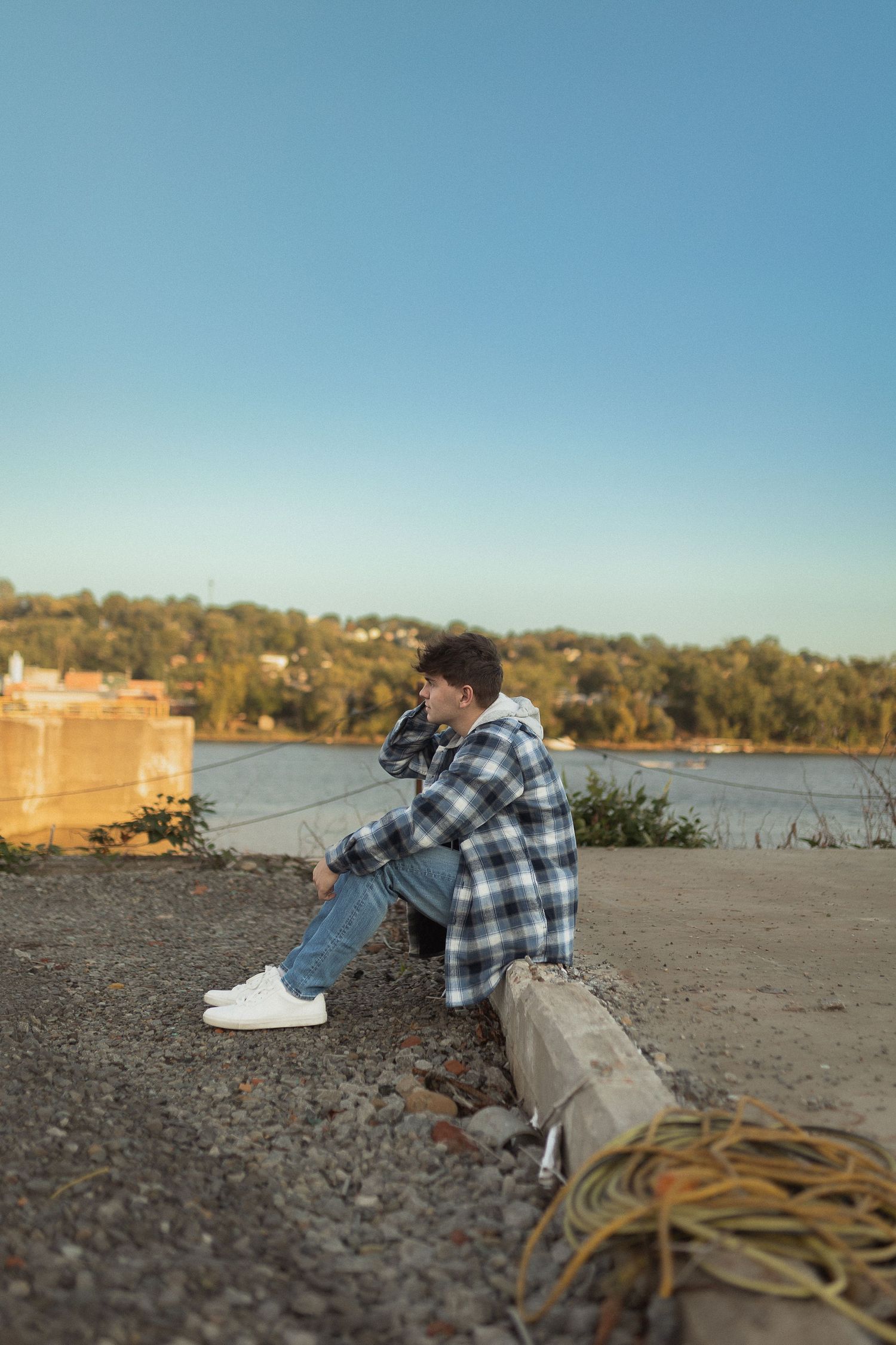 Person in plaid shirt sitting thoughtfully by riverside on cloudy day with bag nearby.