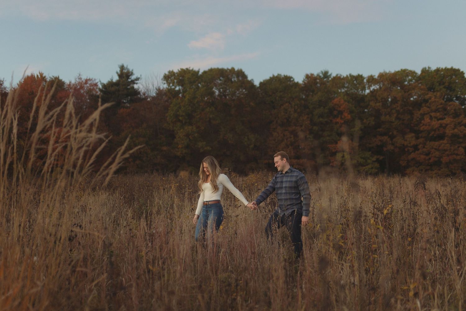 A couple walks hand in hand through a golden field of tall grass during autumn.