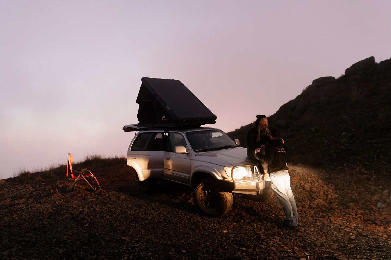 4x4 truck with rooftop tent camping on rocky mountain terrain at dusk with glowing headlights.