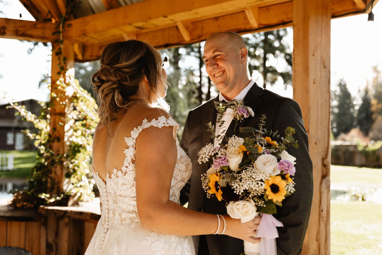 Groom beams with joy as bride in lace gown presents sunflower and rose bouquet under rustic gazebo.