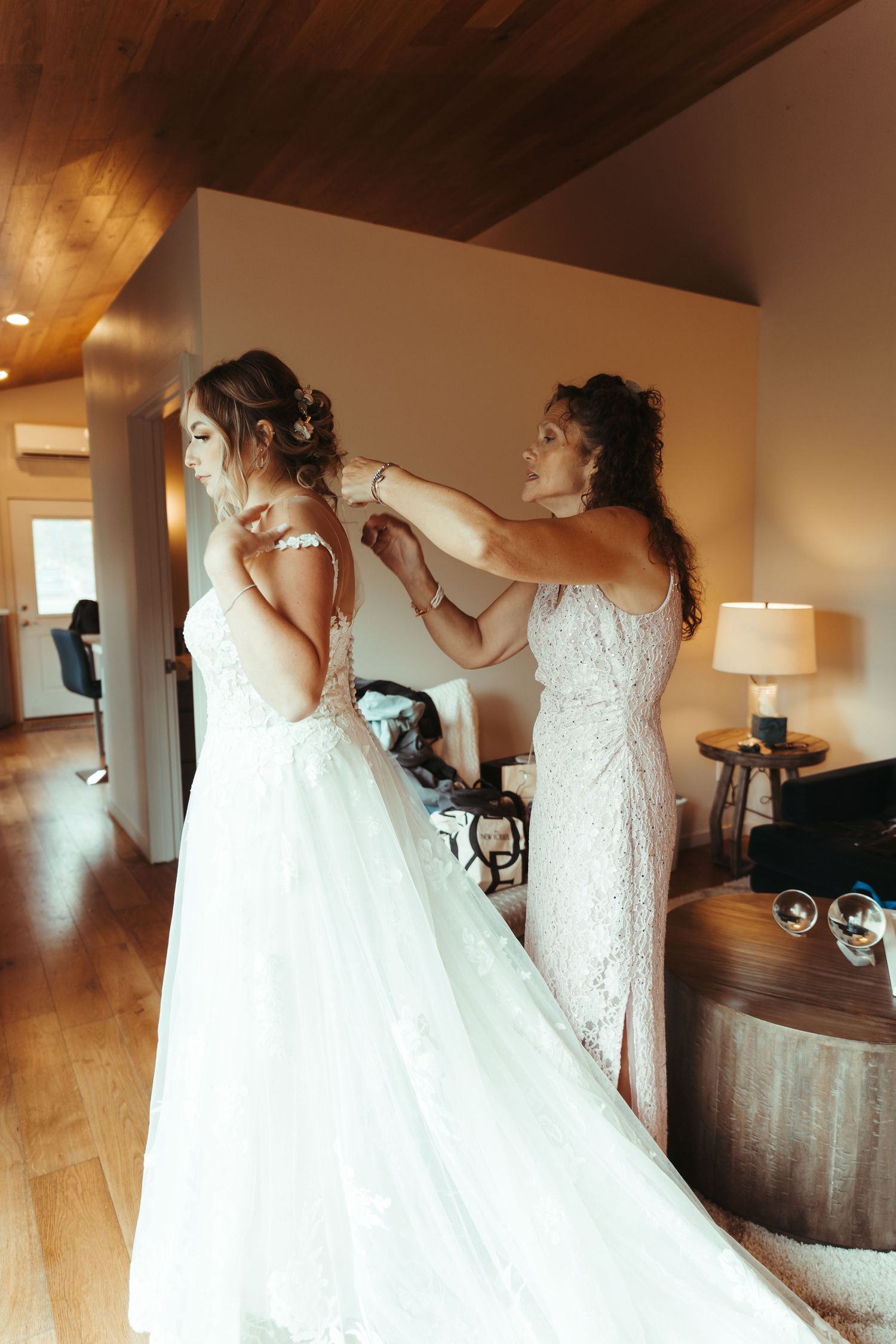 Woman in lace dress fastening back of bride's white wedding gown indoors.