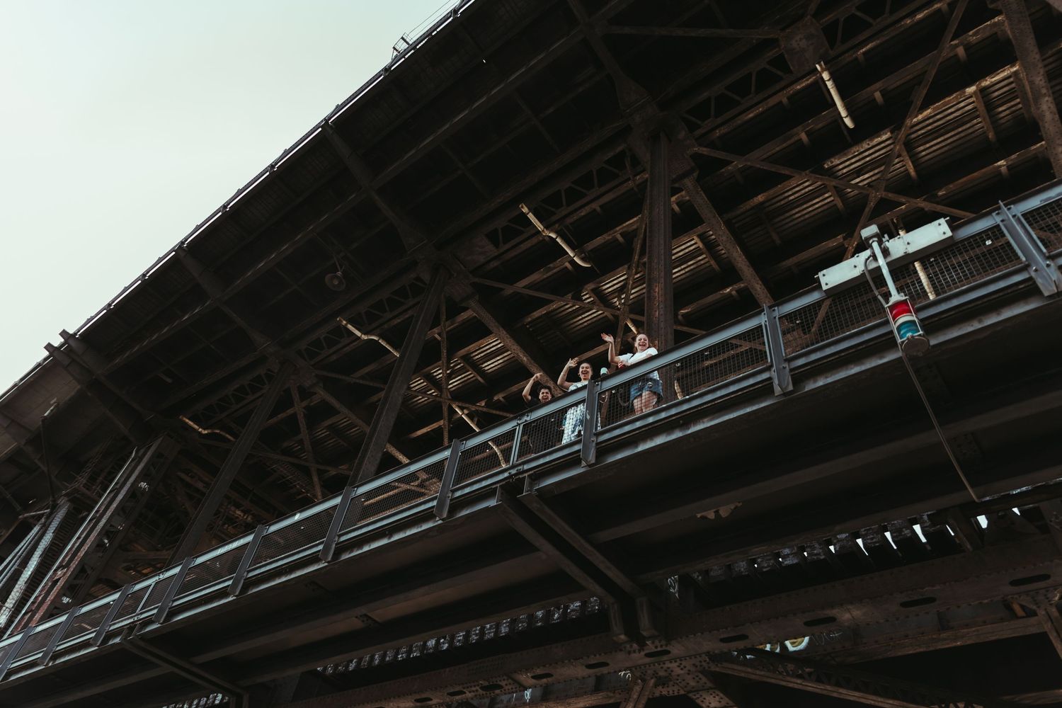 People stand on a metal walkway beneath a large industrial steel bridge structure against a gray sky.