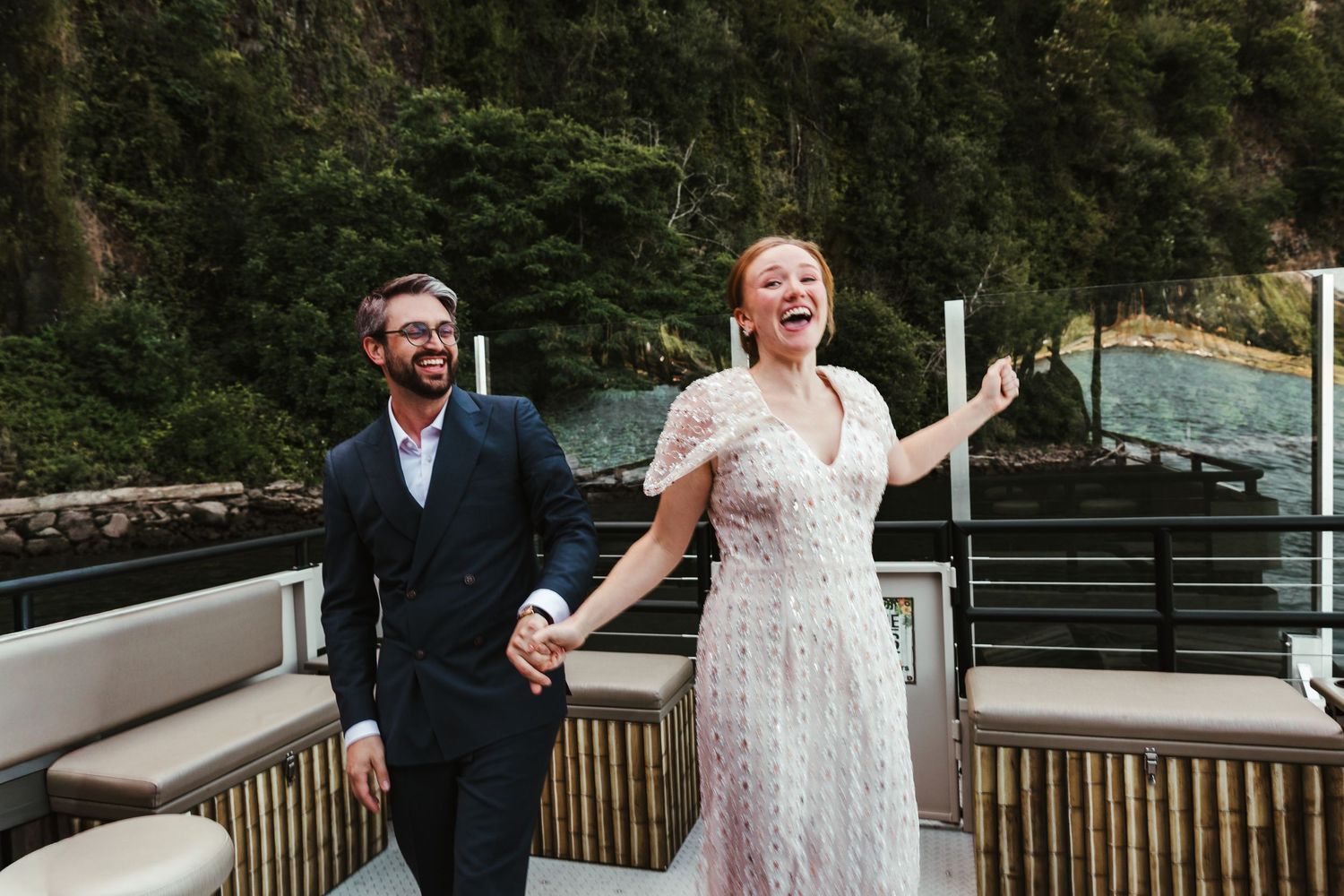 Happy couple in wedding attire laughing and holding hands on a boat deck with lush green trees in the background.