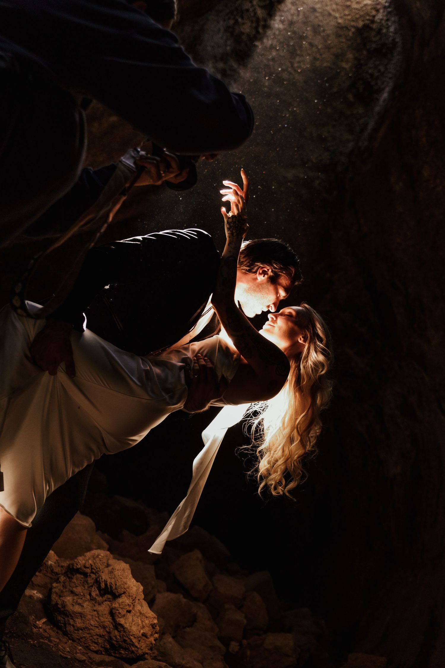 Couple in a dramatic dip kiss inside a dark rocky cave, her long blonde hair and white dress lit by a beam of light.