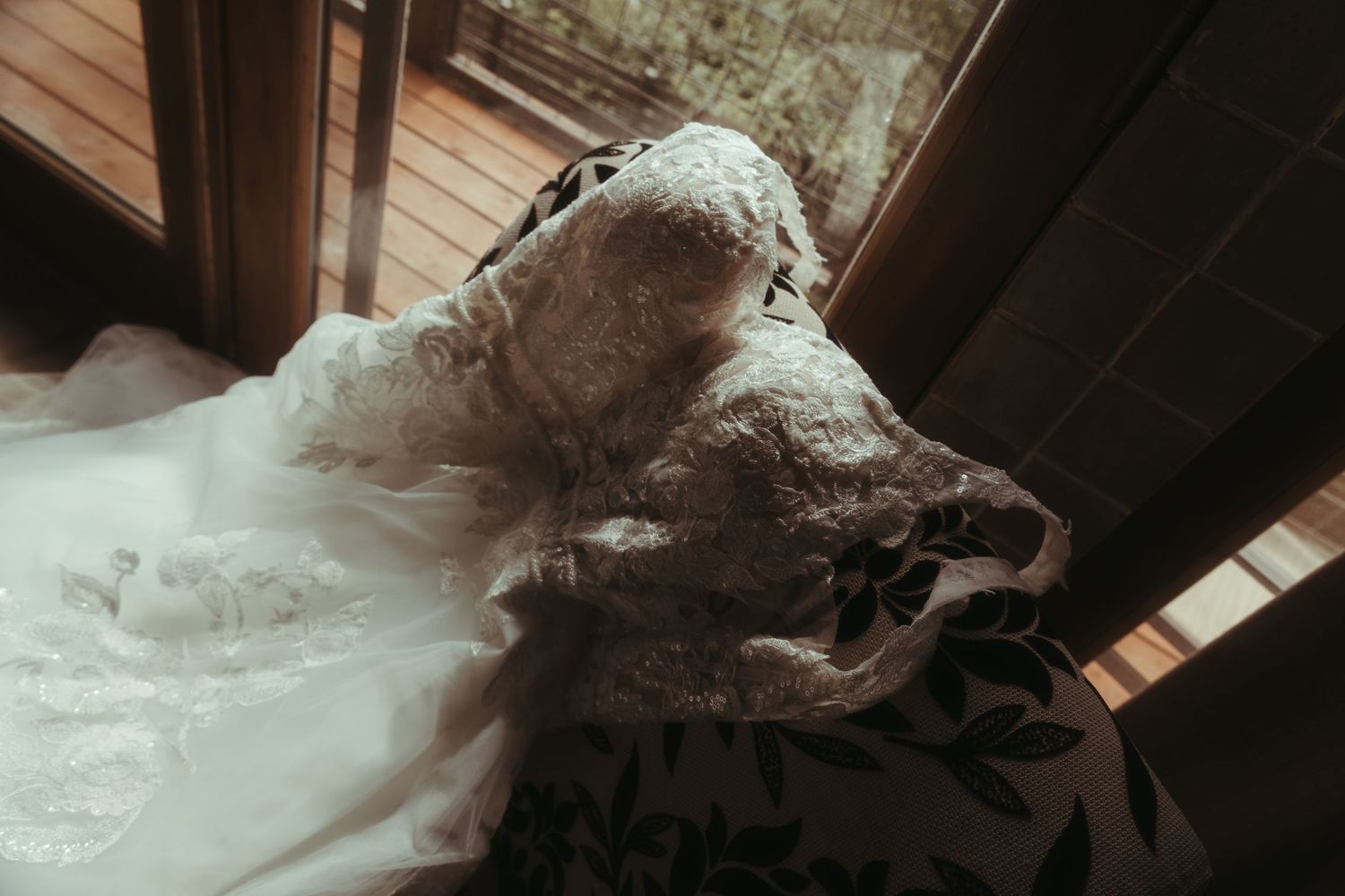 A delicate white lace wedding dress draped over a floral chair near a sunlit window.