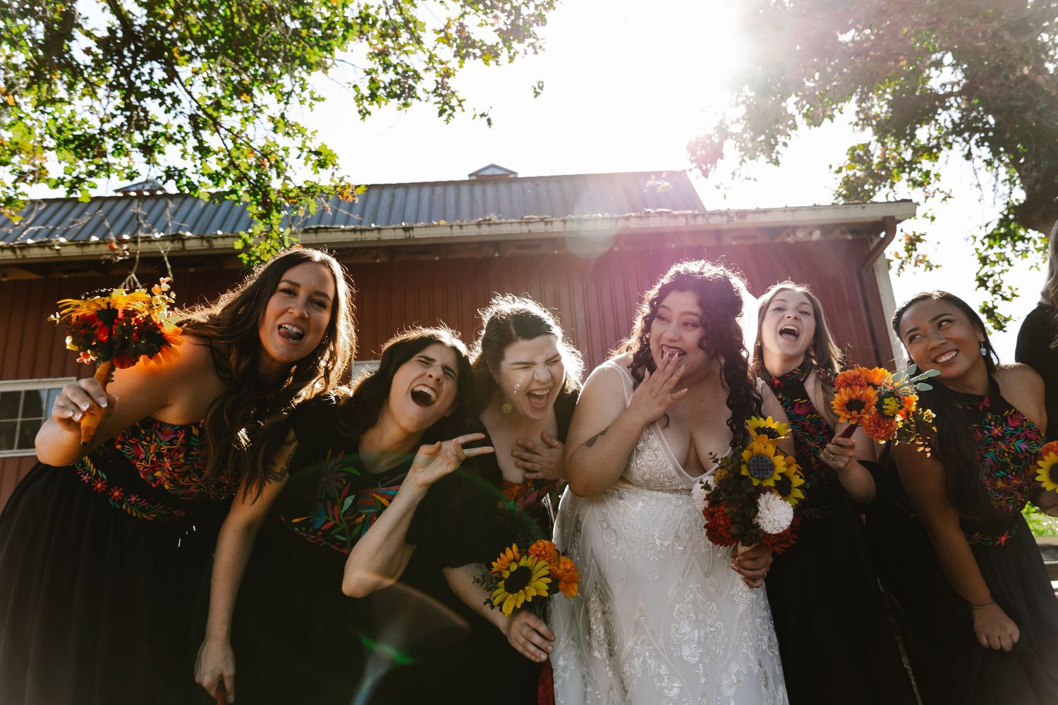 Bride in a white gown laughs with bridesmaids in black dresses holding sunflower bouquets outside a rustic red barn on a sunny day.