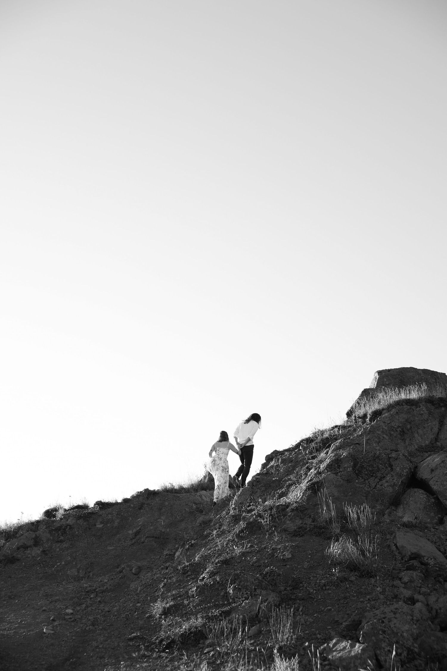 Black and white photo of a couple holding hands while climbing a steep rocky hillside against a bright, clear sky.