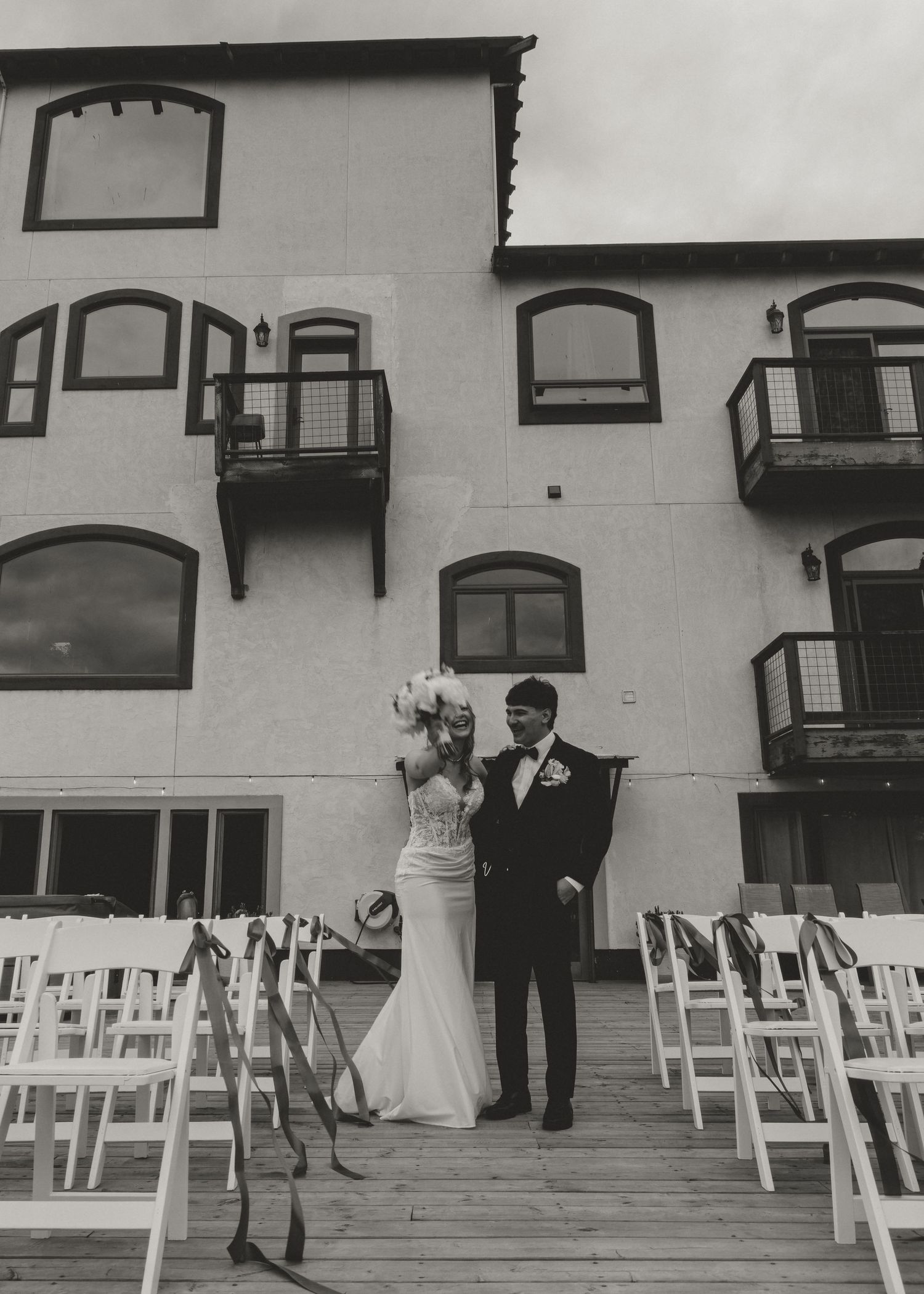 Black and white photo of a bride and groom standing between rows of white chairs at an outdoor wedding ceremony venue.