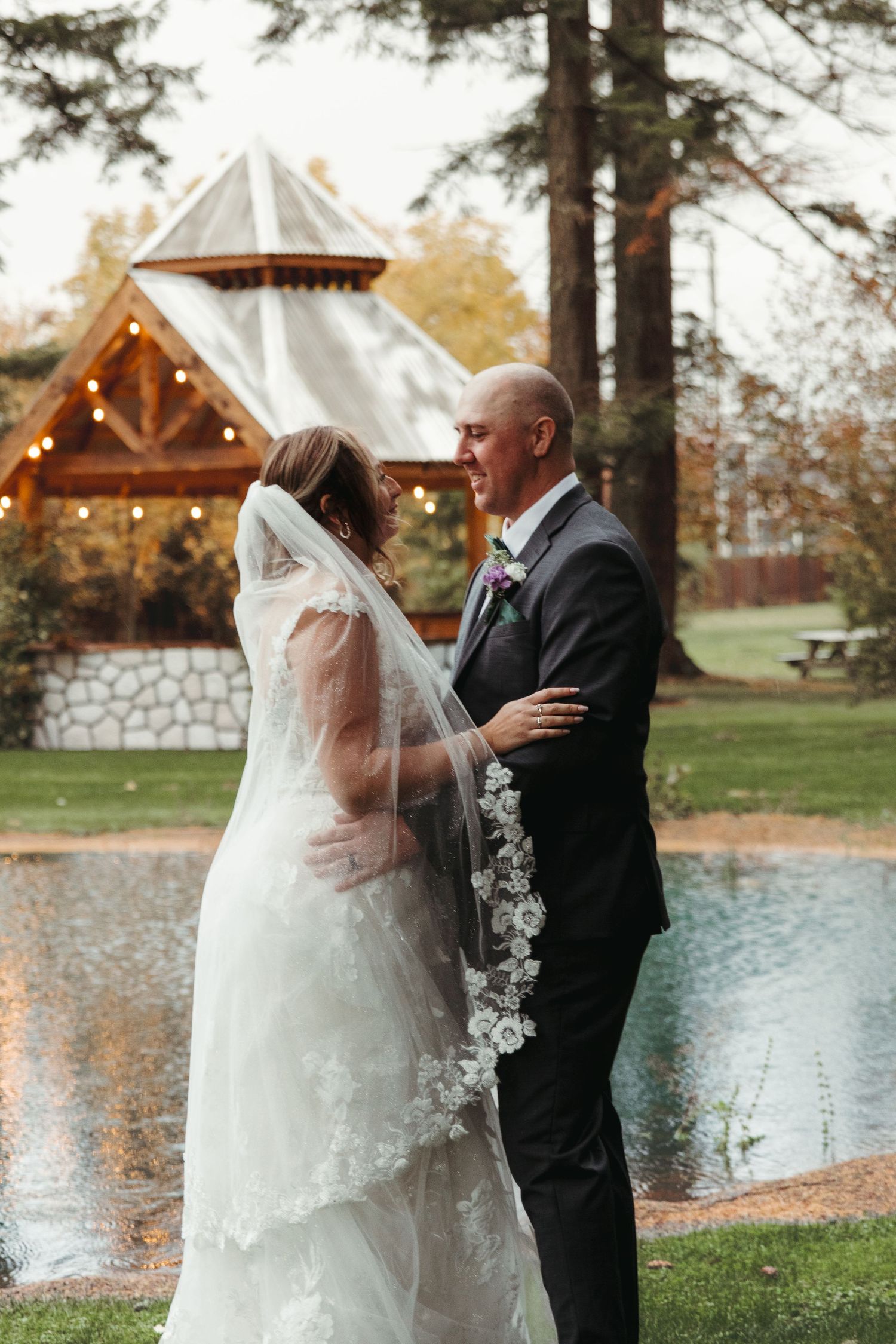 Bride and groom share a romantic moment by a pond at an outdoor wedding venue with string lights.
