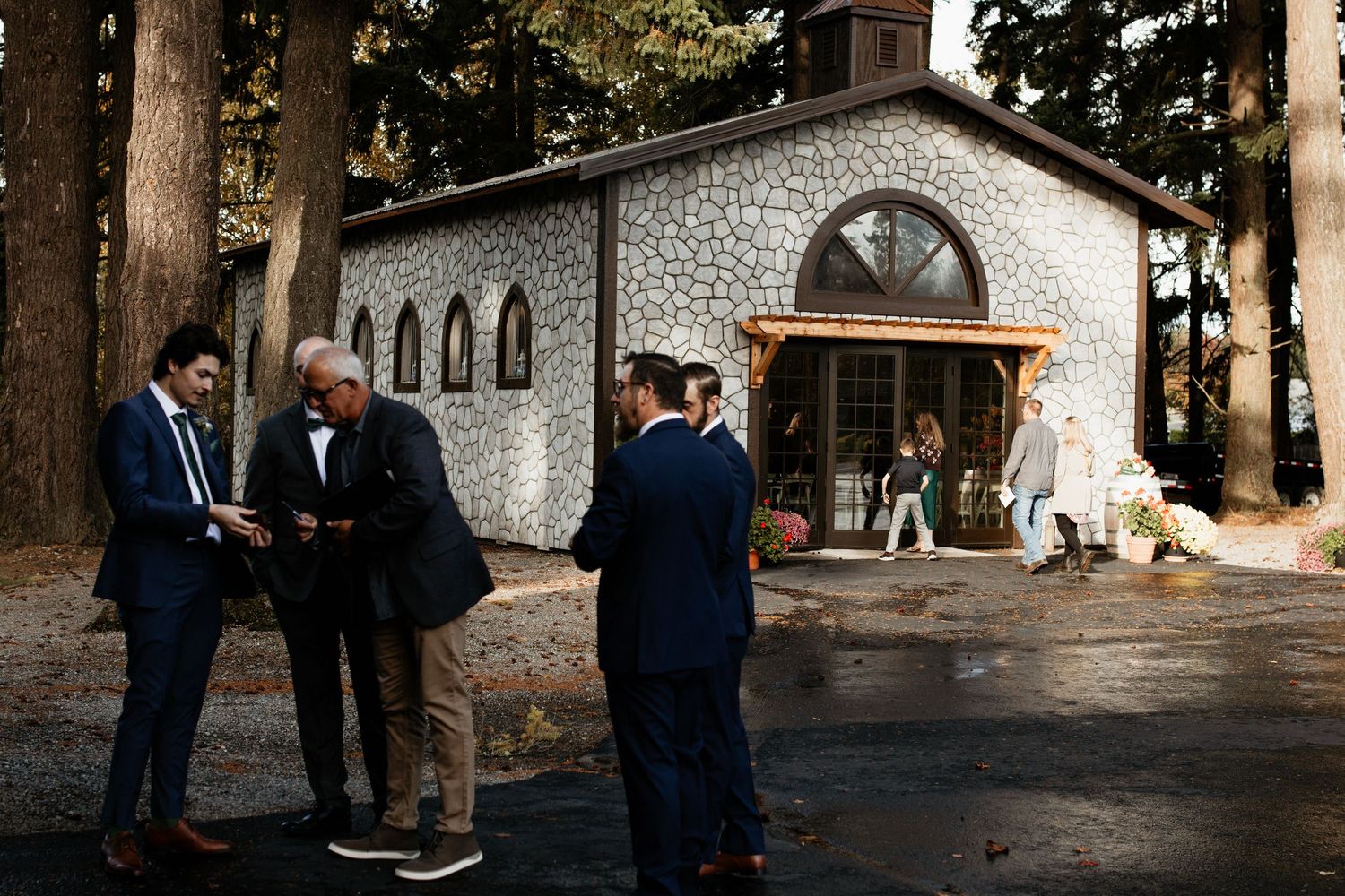 Wedding guests mingle outside a rustic stone chapel amid towering trees before the ceremony.