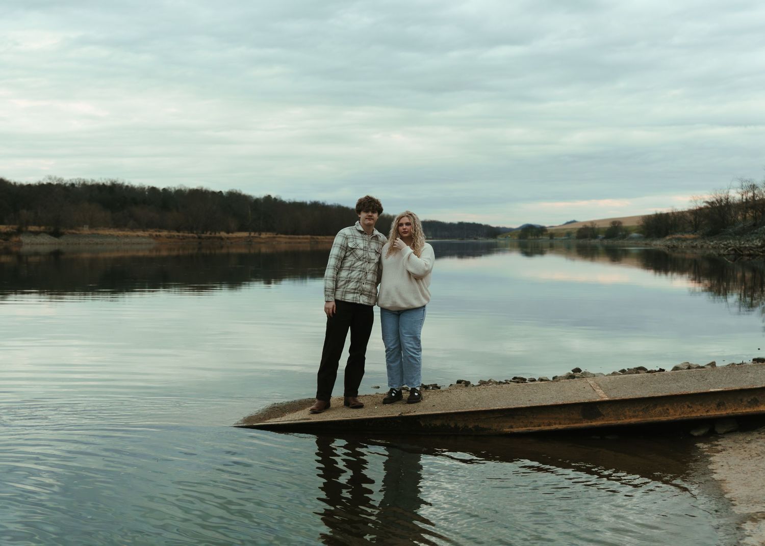 A couple standing on a wooden dock over a calm reflective lake under a cloudy sky in autumn.