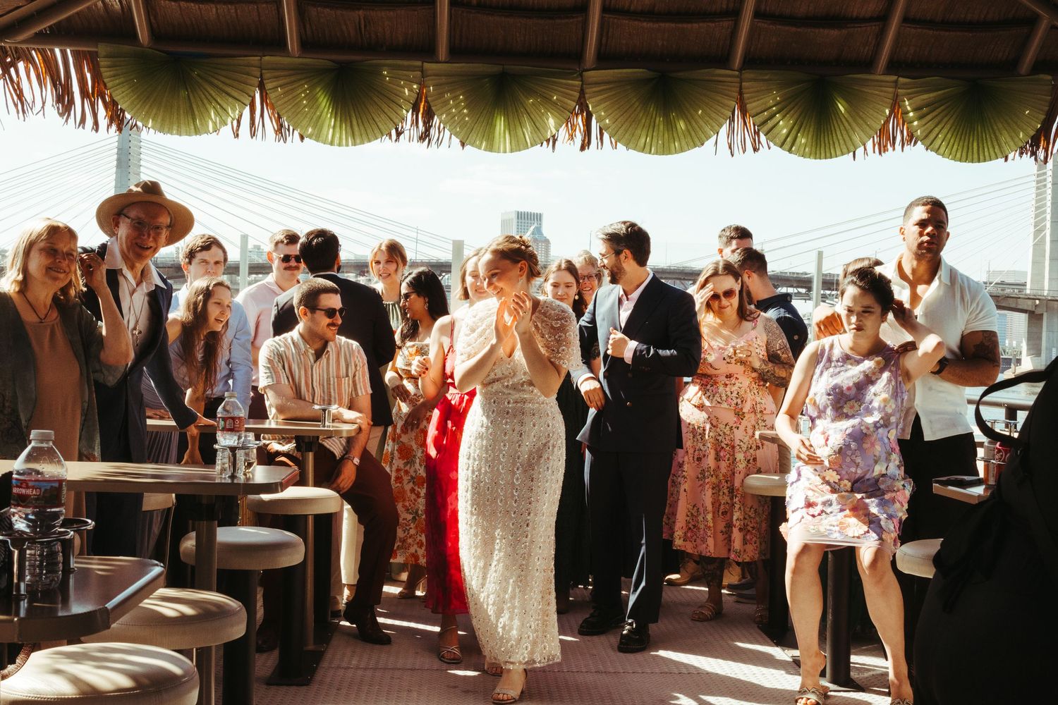 Guests celebrate at a rooftop party under palm fronds, with a woman in a sequin dress at the center.