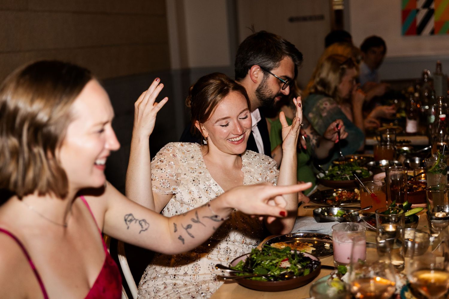 Women laughing and pointing at a long dinner table filled with food and drinks at a lively restaurant gathering.