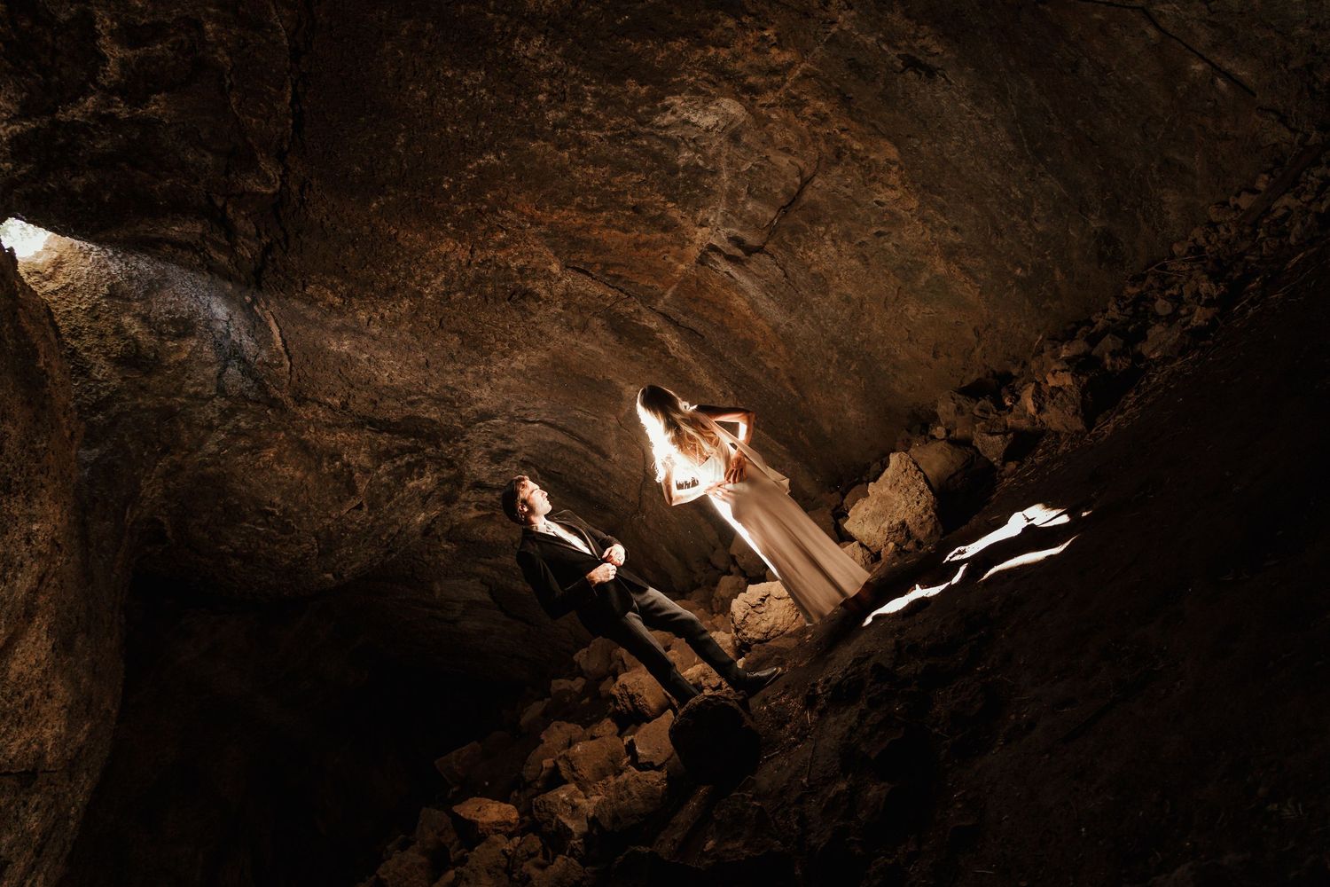 Couple in elegant attire poses dramatically inside a dark cave with rocky walls and atmospheric lighting.