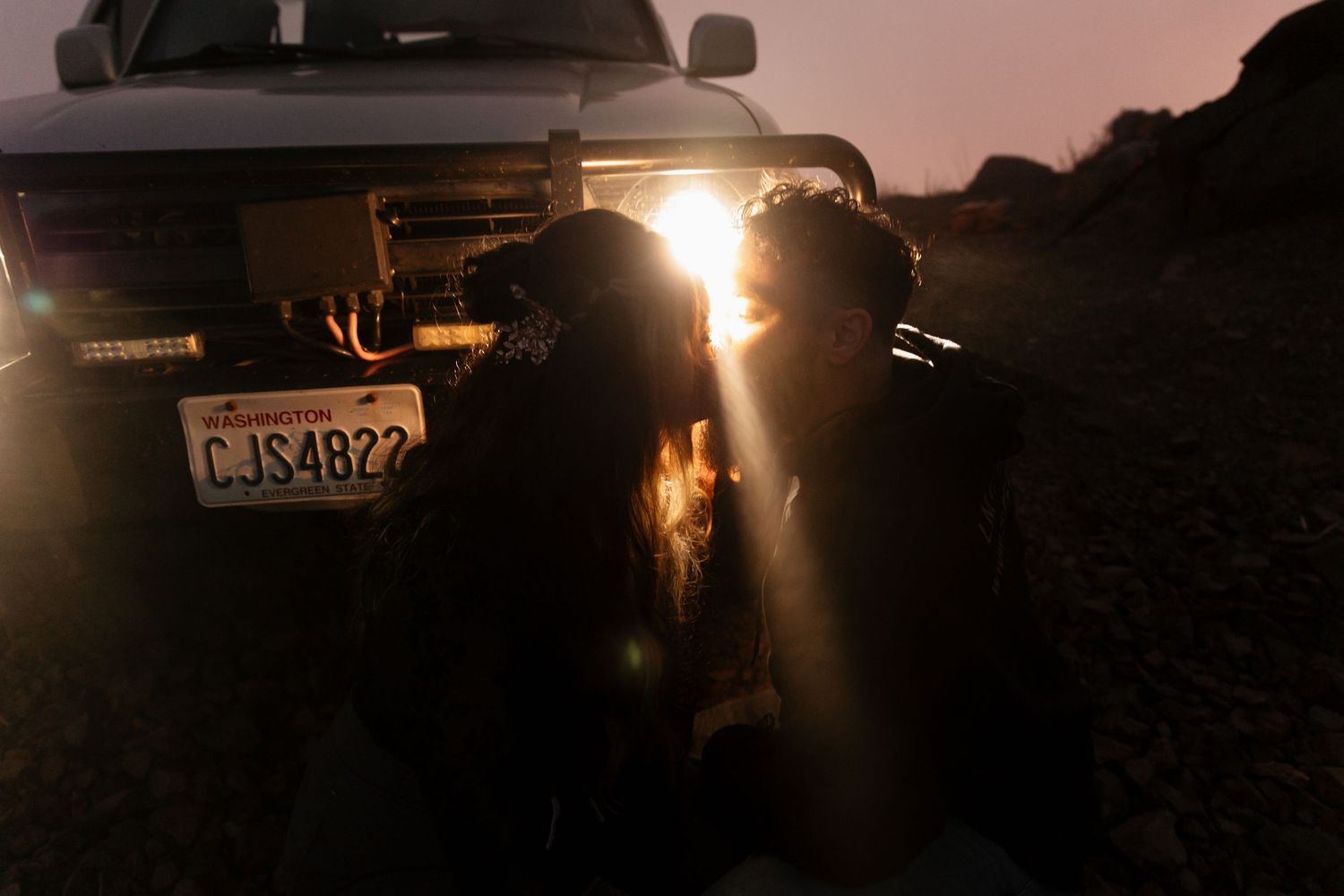 Couple sharing a romantic kiss in front of illuminated truck headlights at dusk with Washington license plate.