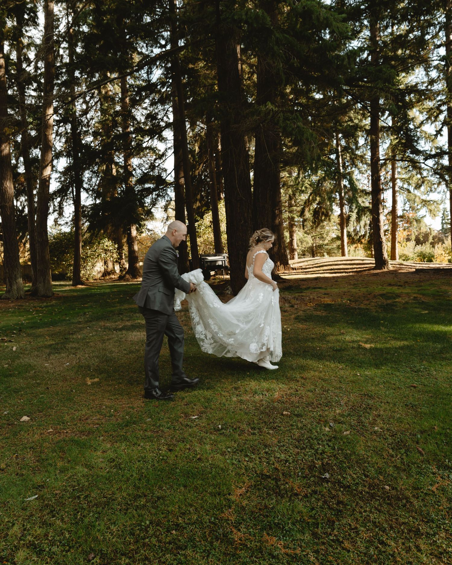 Groom helps carry bride's white wedding gown train across grassy area surrounded by tall pine trees.