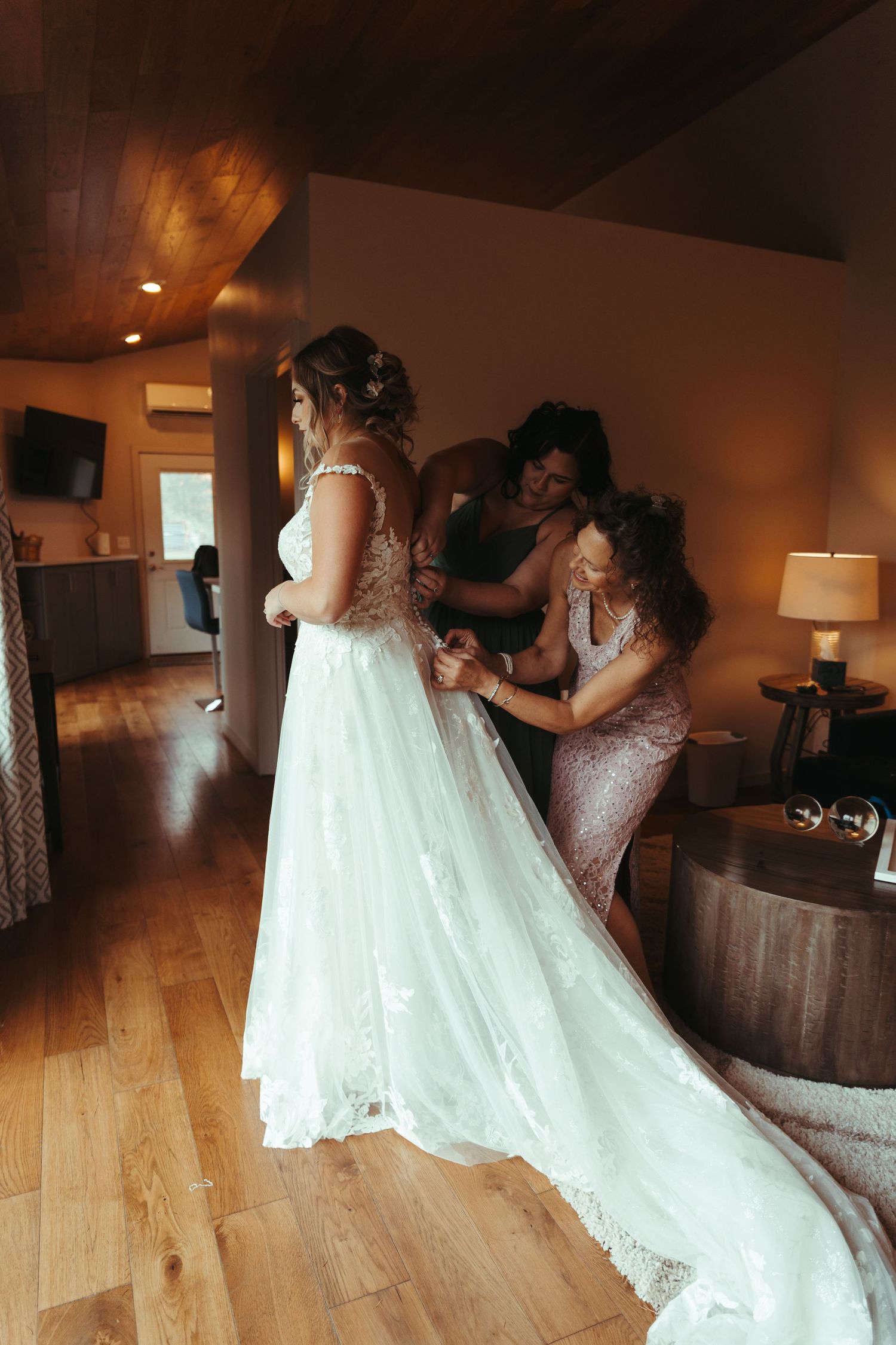 Two women assisting bride with back of flowing white lace wedding gown in warm interior.