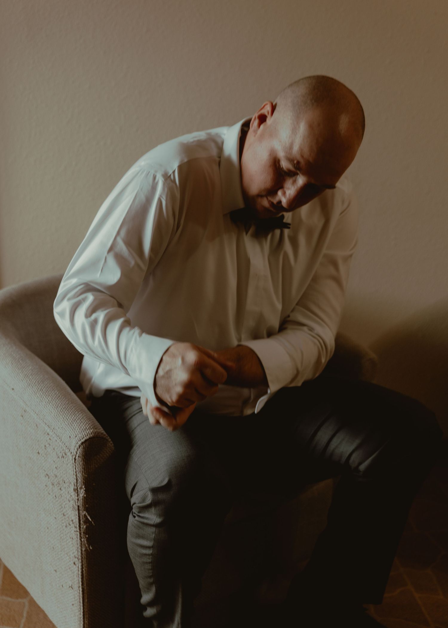 A bald man in a white dress shirt and bow tie sits in a chair adjusting his cufflinks.