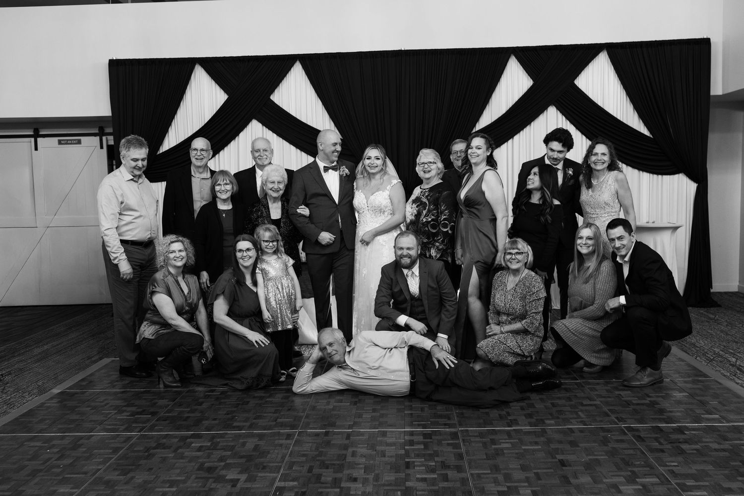 Black and white photo of a large wedding party group posing in front of draped black and white backdrop.