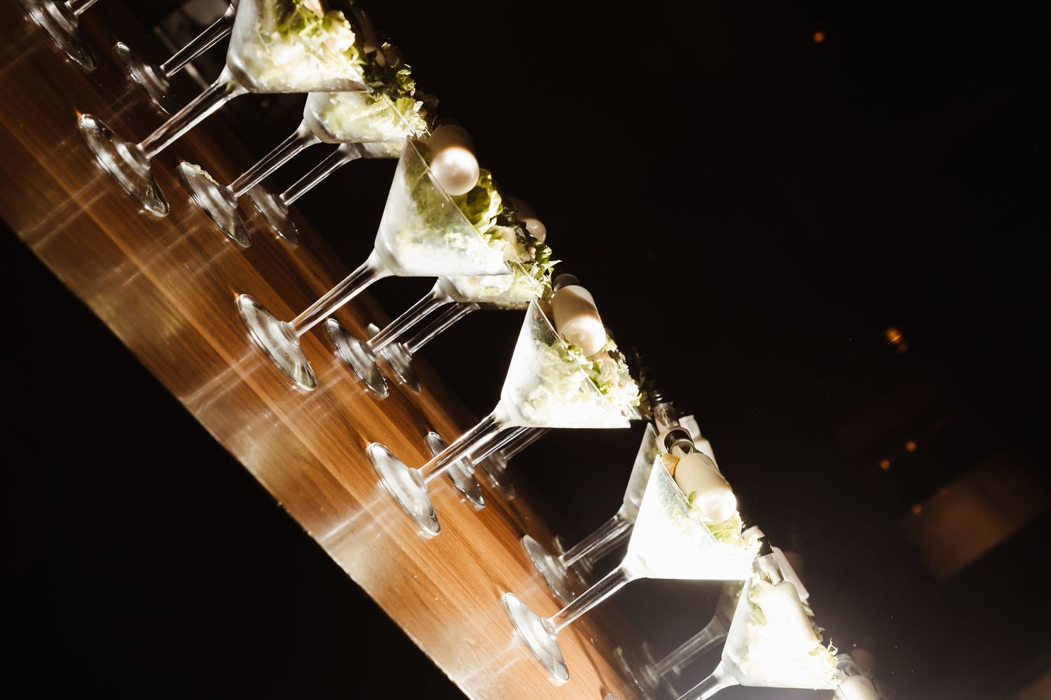 A row of elegant martini glasses filled with white floral arrangements on a illuminated wooden table at an event.