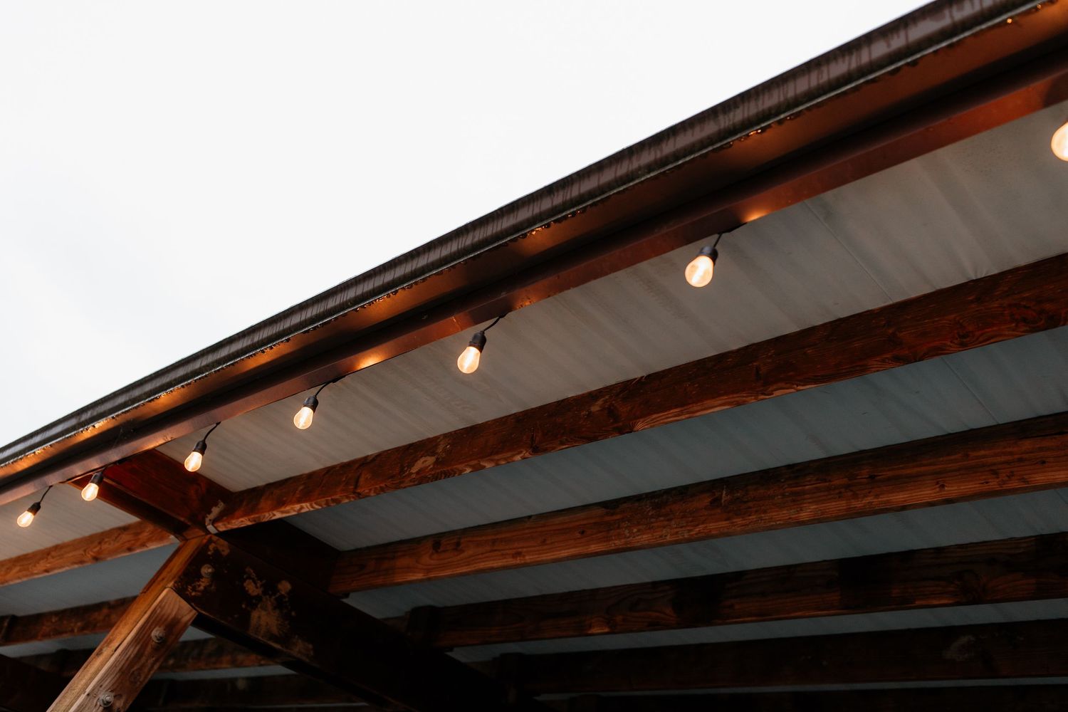 String of globe lights hanging beneath wooden beams of an outdoor wedding venue canopy.