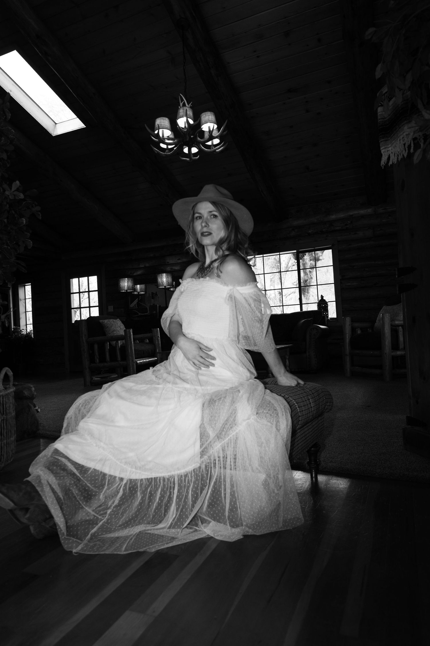 Black and white photo of a woman in a white dress and hat sitting elegantly in a rustic log cabin interior.