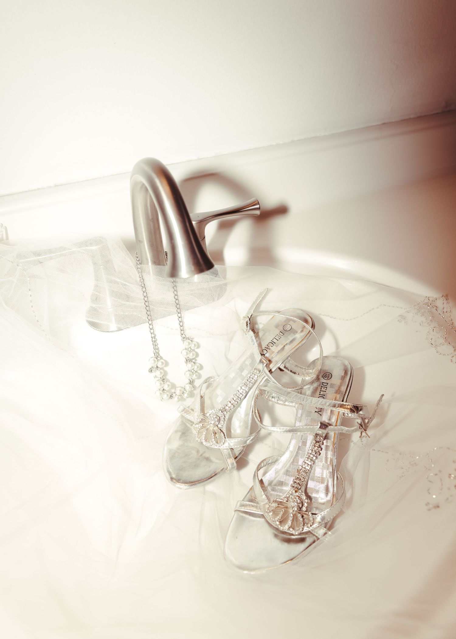 Silver bridal sandals and pearl necklace displayed elegantly on white fabric near a brushed nickel bathroom faucet.